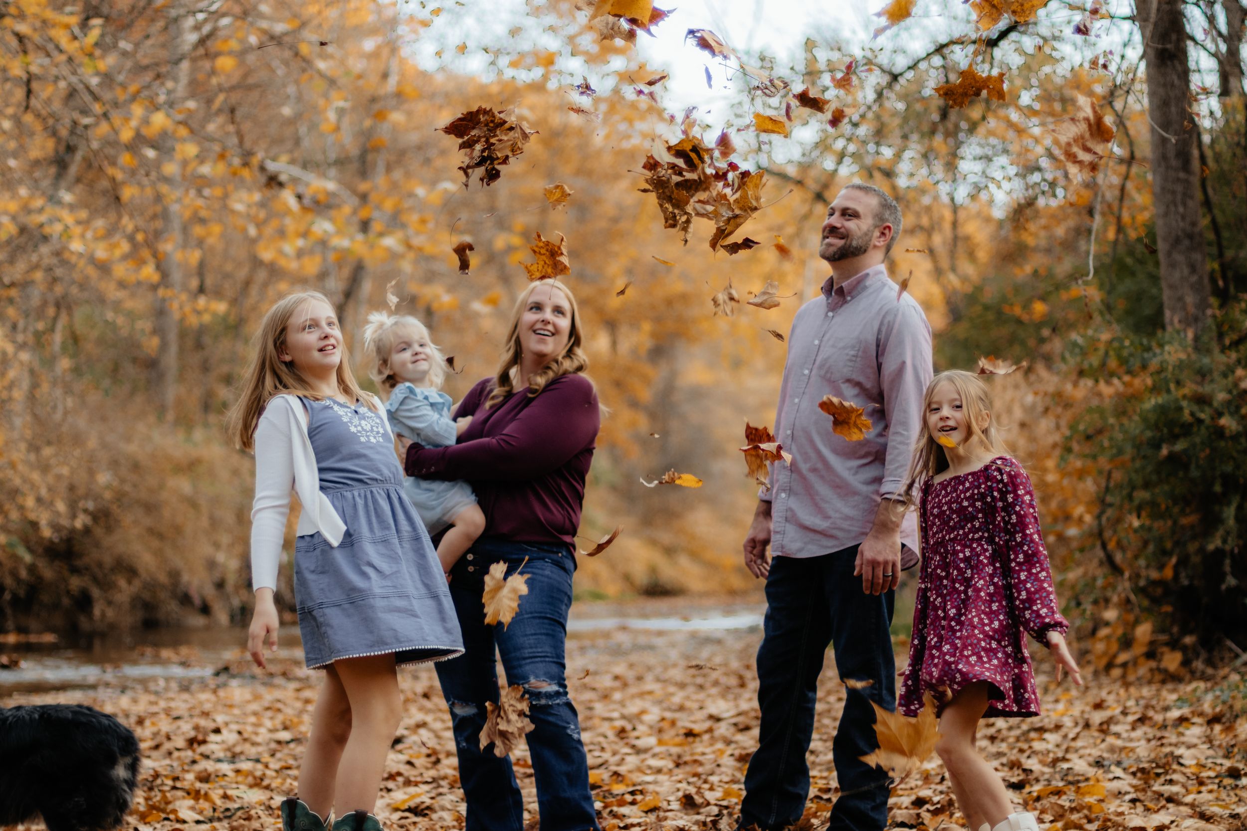Family members laugh and play in fallen autumn leaves during an outdoor photo session.