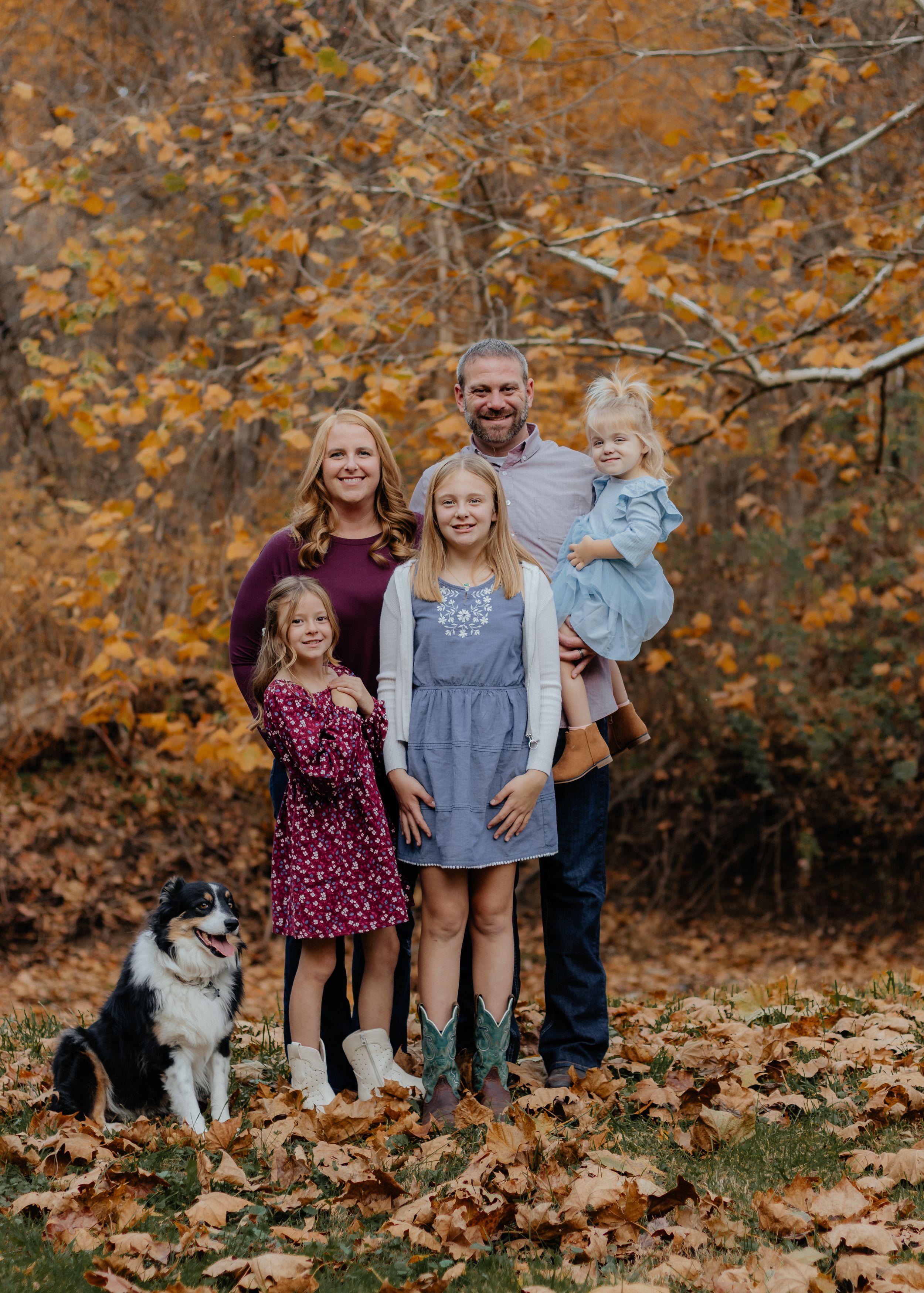 Family portrait with black and white dog against fall foliage backdrop.