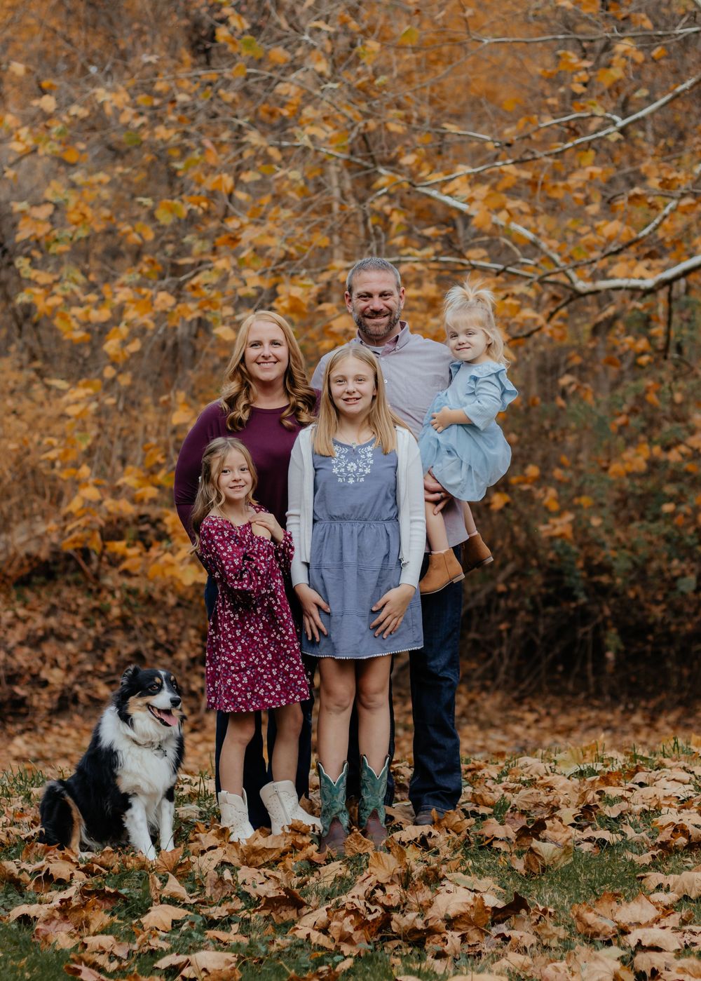 Family portrait with black and white dog against fall foliage backdrop.