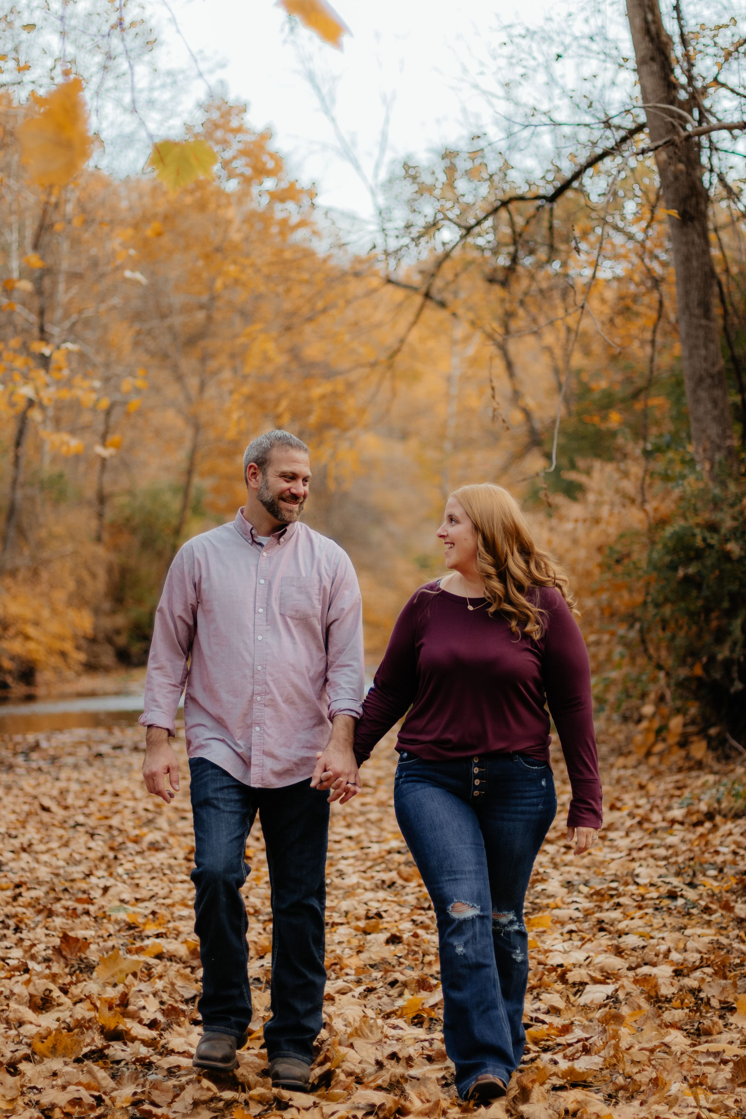 Couple walking hand in hand through fallen leaves during autumn photo session.