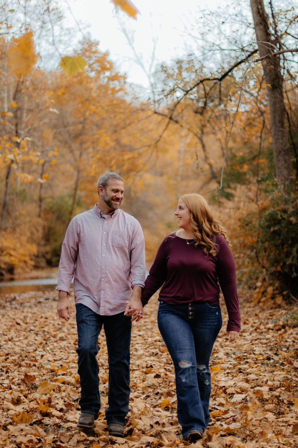 Couple walking hand in hand through fallen leaves during autumn photo session.