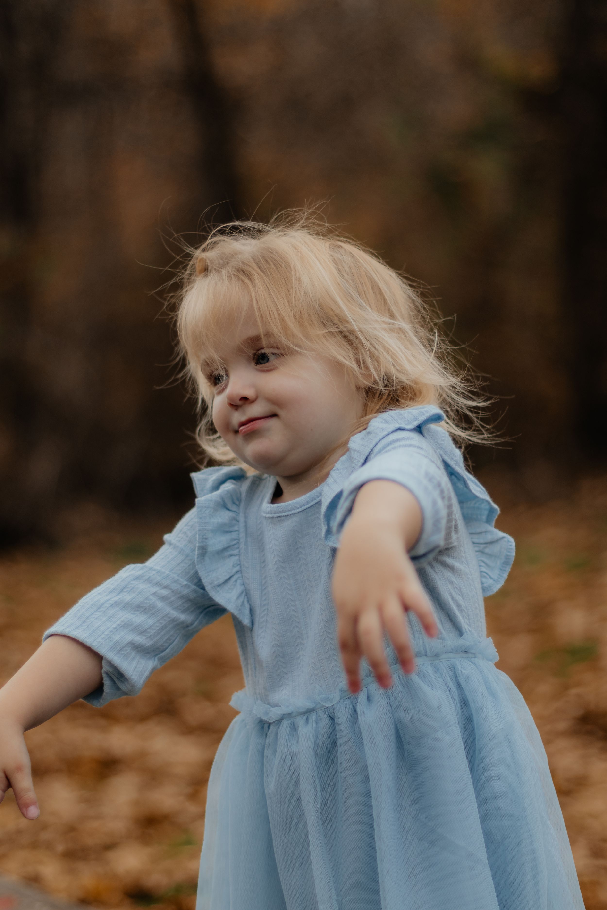 Young child in light blue dress plays joyfully among autumn leaves during outdoor photoshoot.