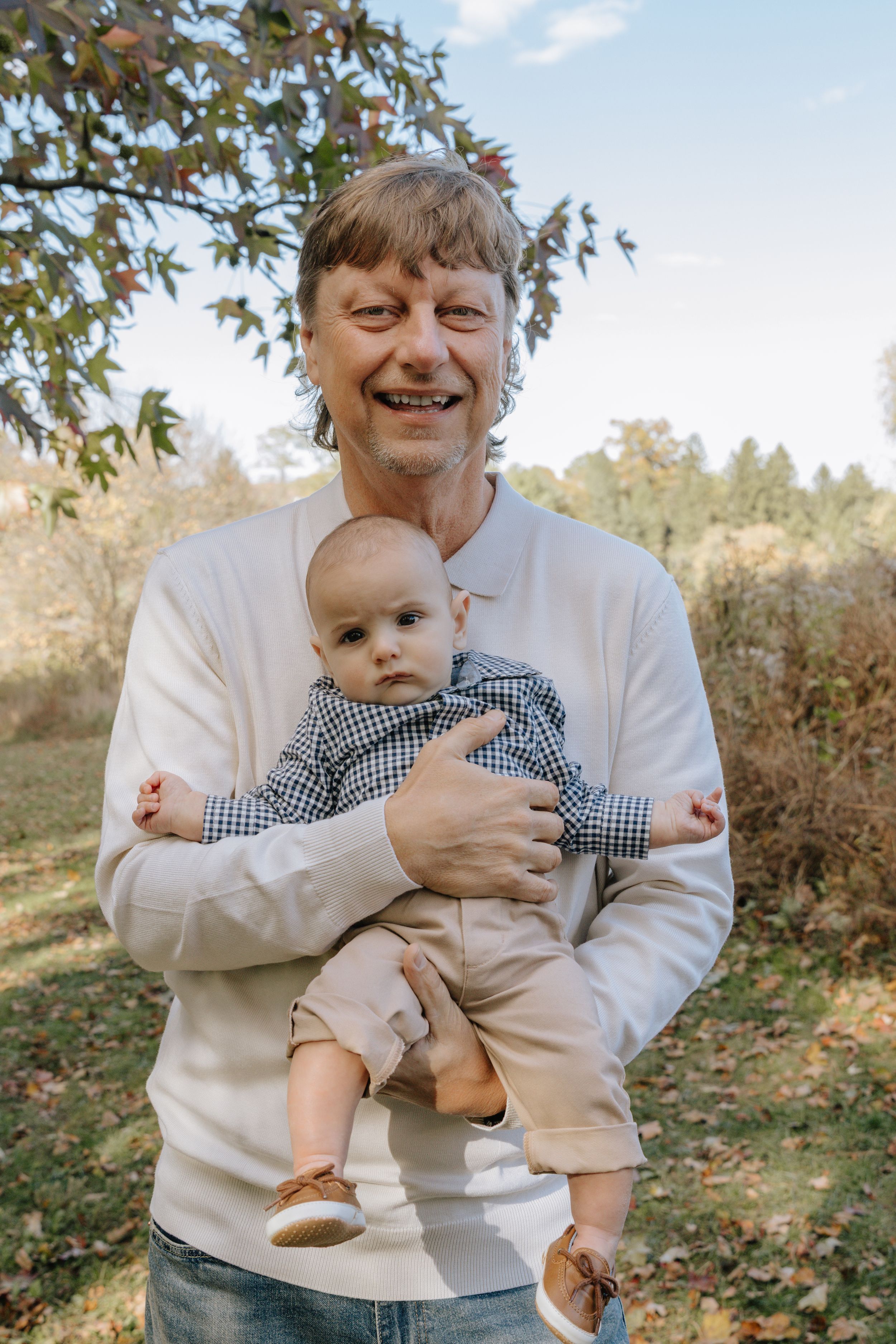 Baby in khaki outfit being held outdoors during autumn photoshoot.