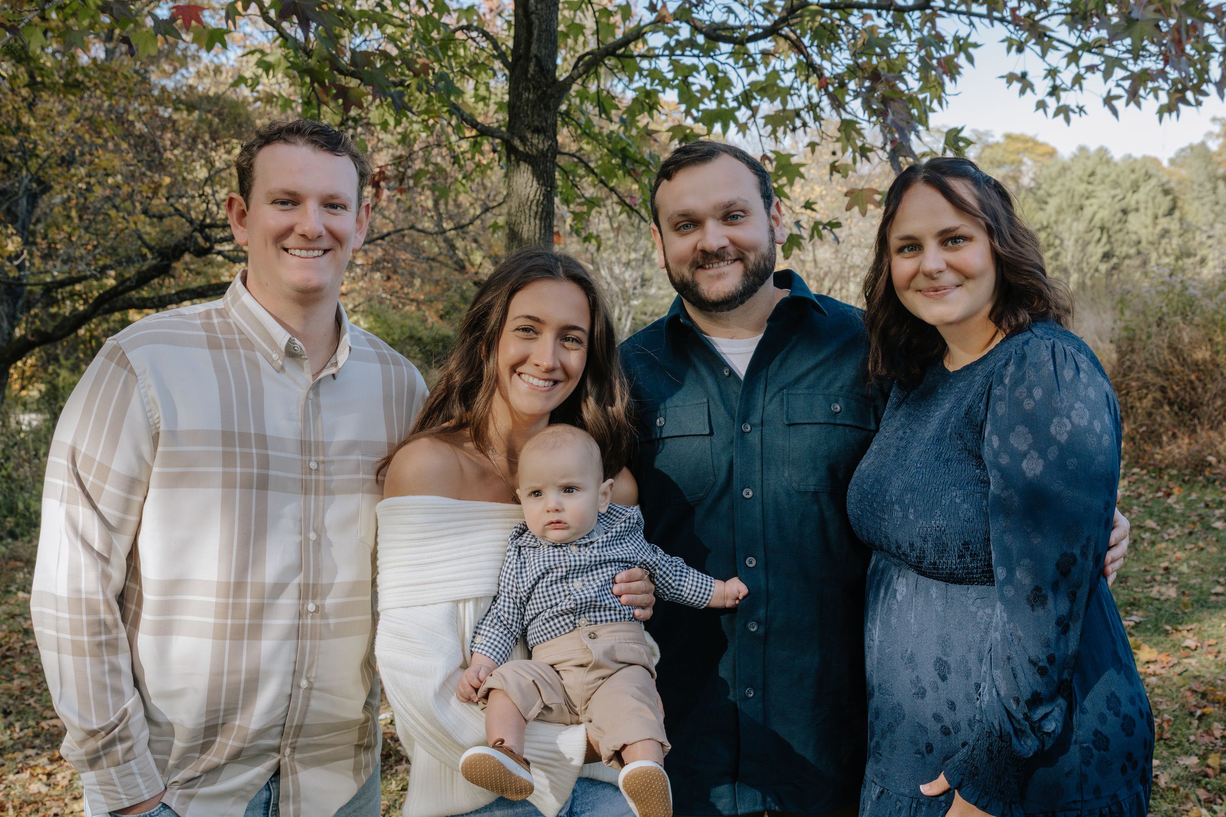 Family group stands together smiling during a fall outdoor photo session with trees in background.