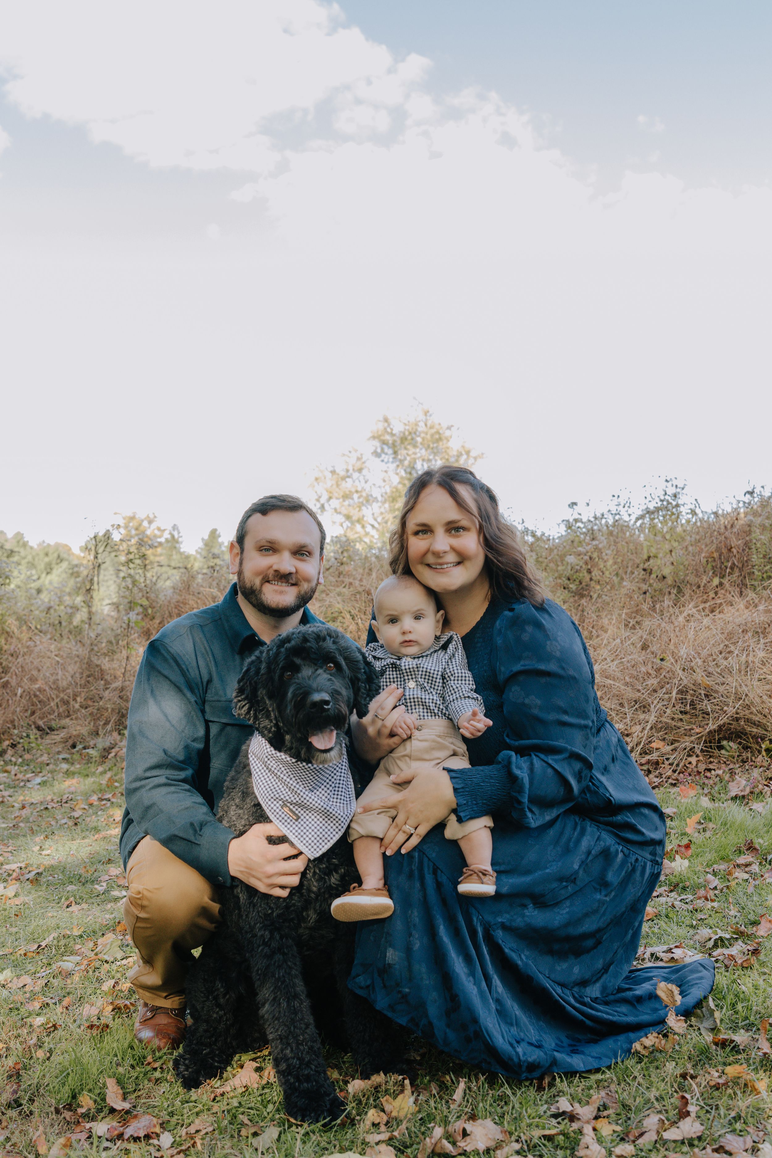 Family sits on grassy hill with their black dog during outdoor photo session.