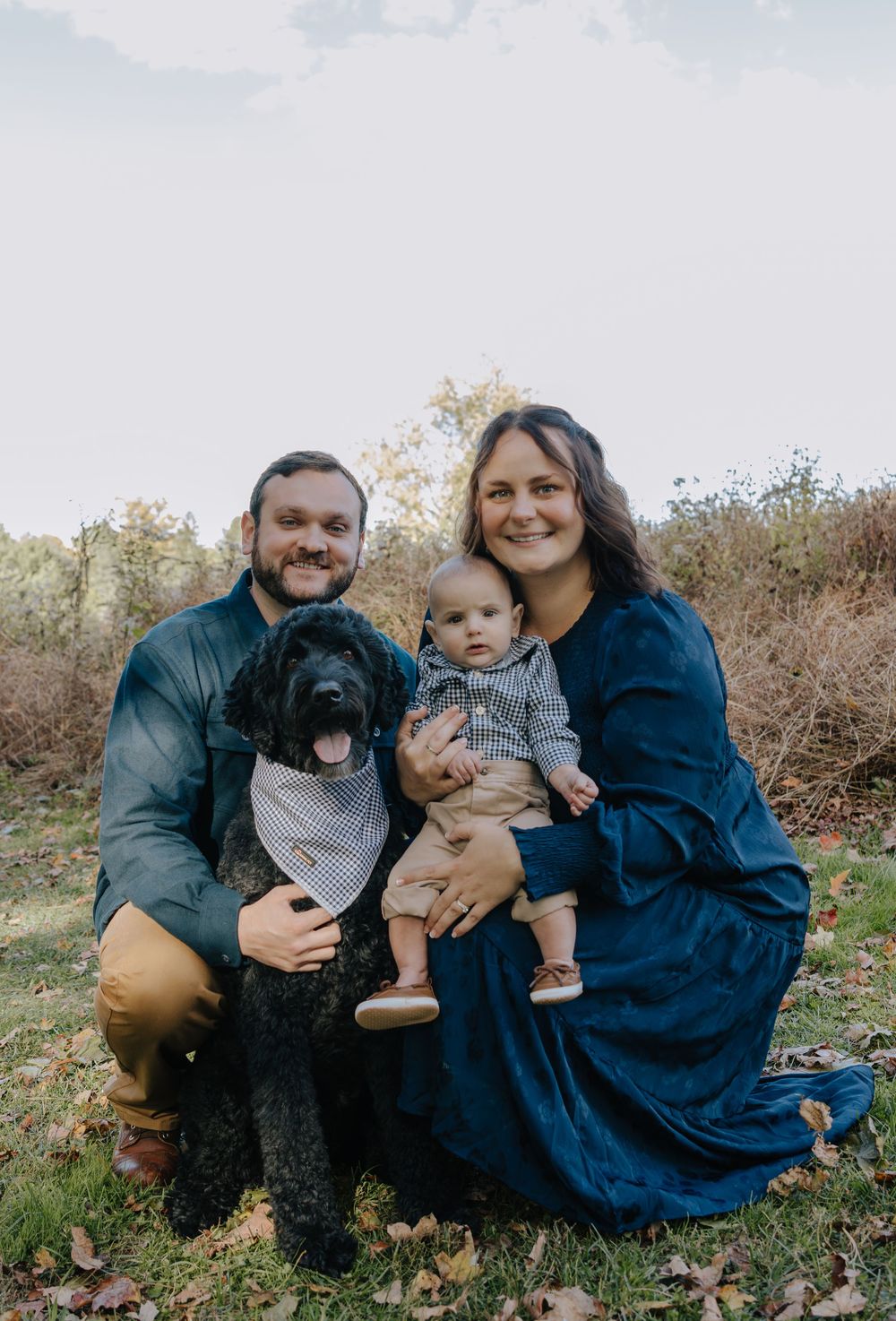 Family group relaxes on grass with their black dog during casual outdoor photoshoot.