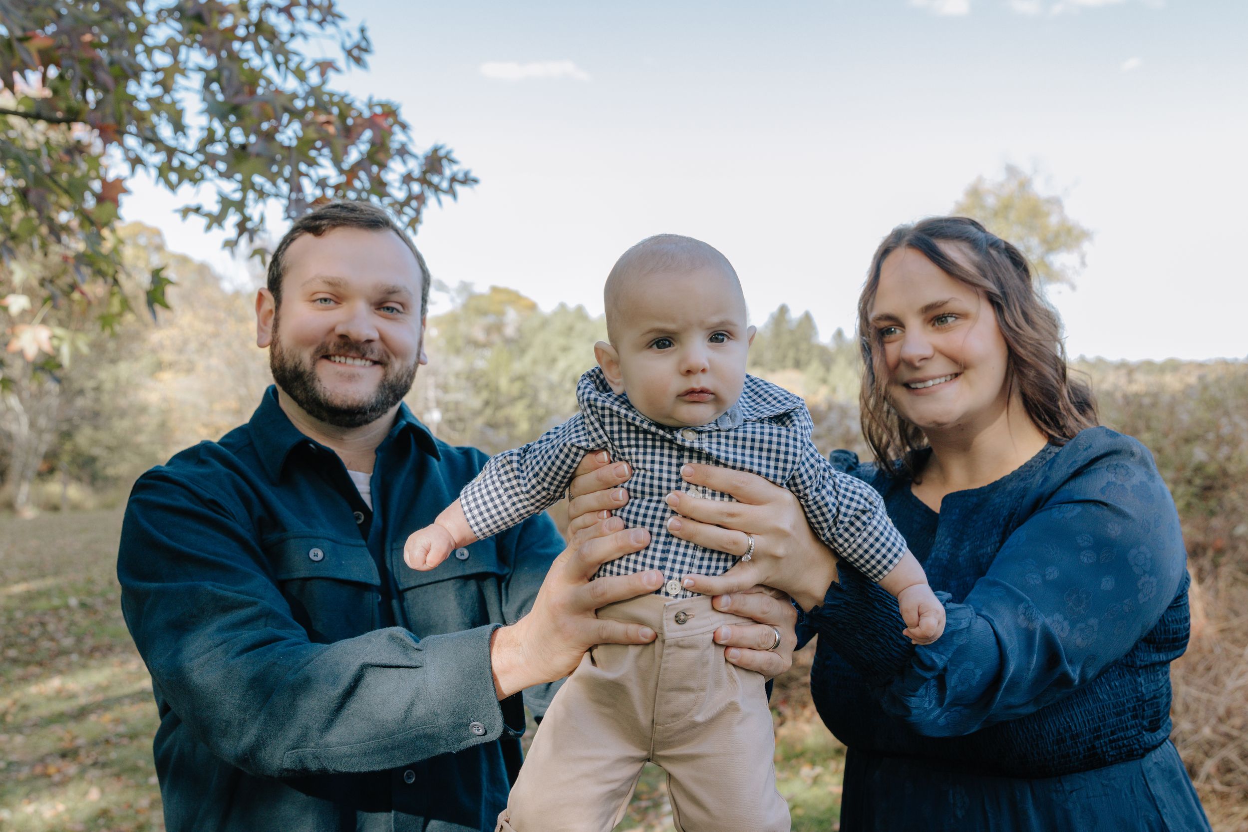 Candid family portrait showing parents embracing their infant child outdoors.