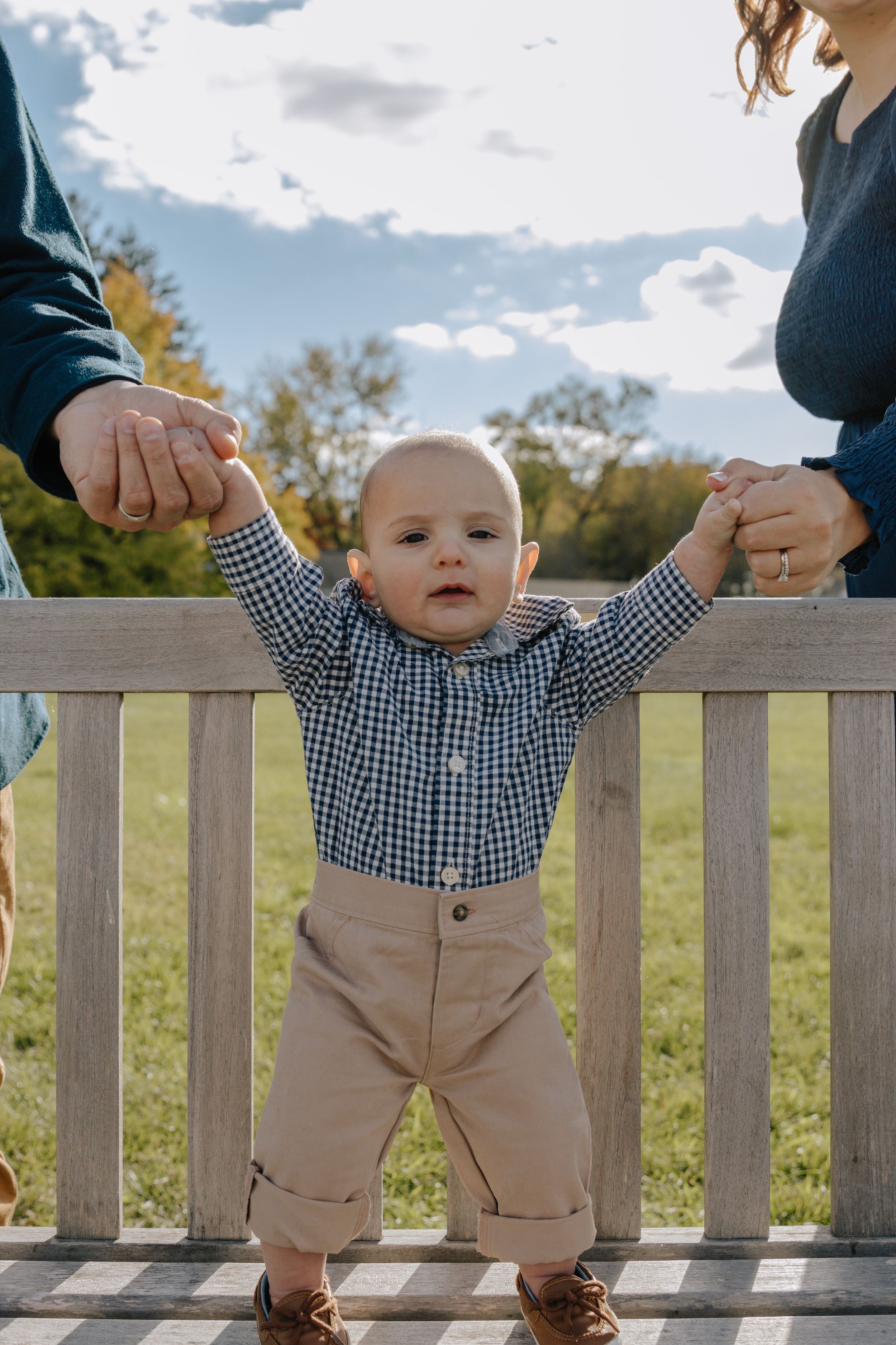 Baby in gingham shirt and khaki pants holding wooden fence rail outdoors.