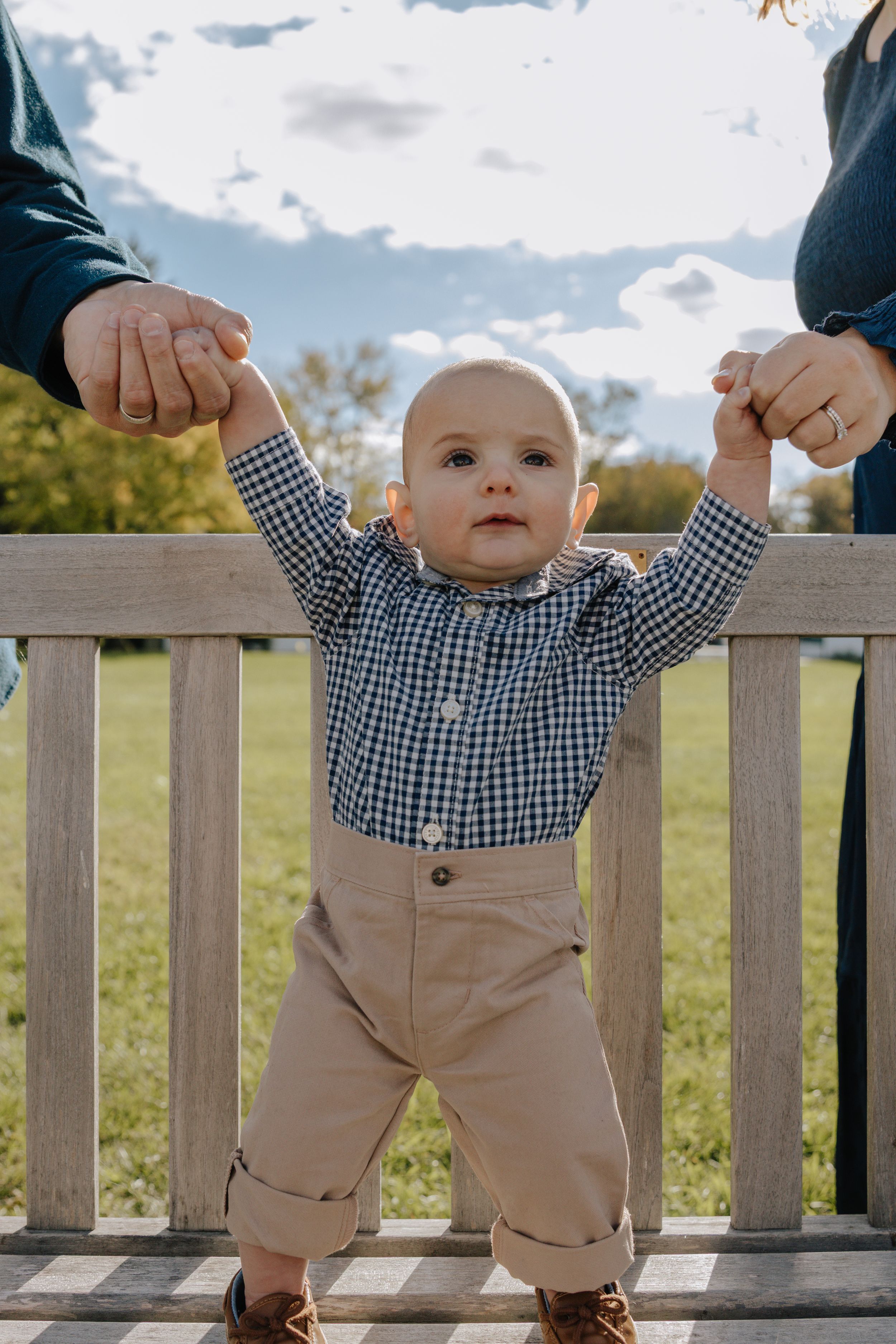Toddler in checkered shirt and khaki pants reaching up while standing by fence.