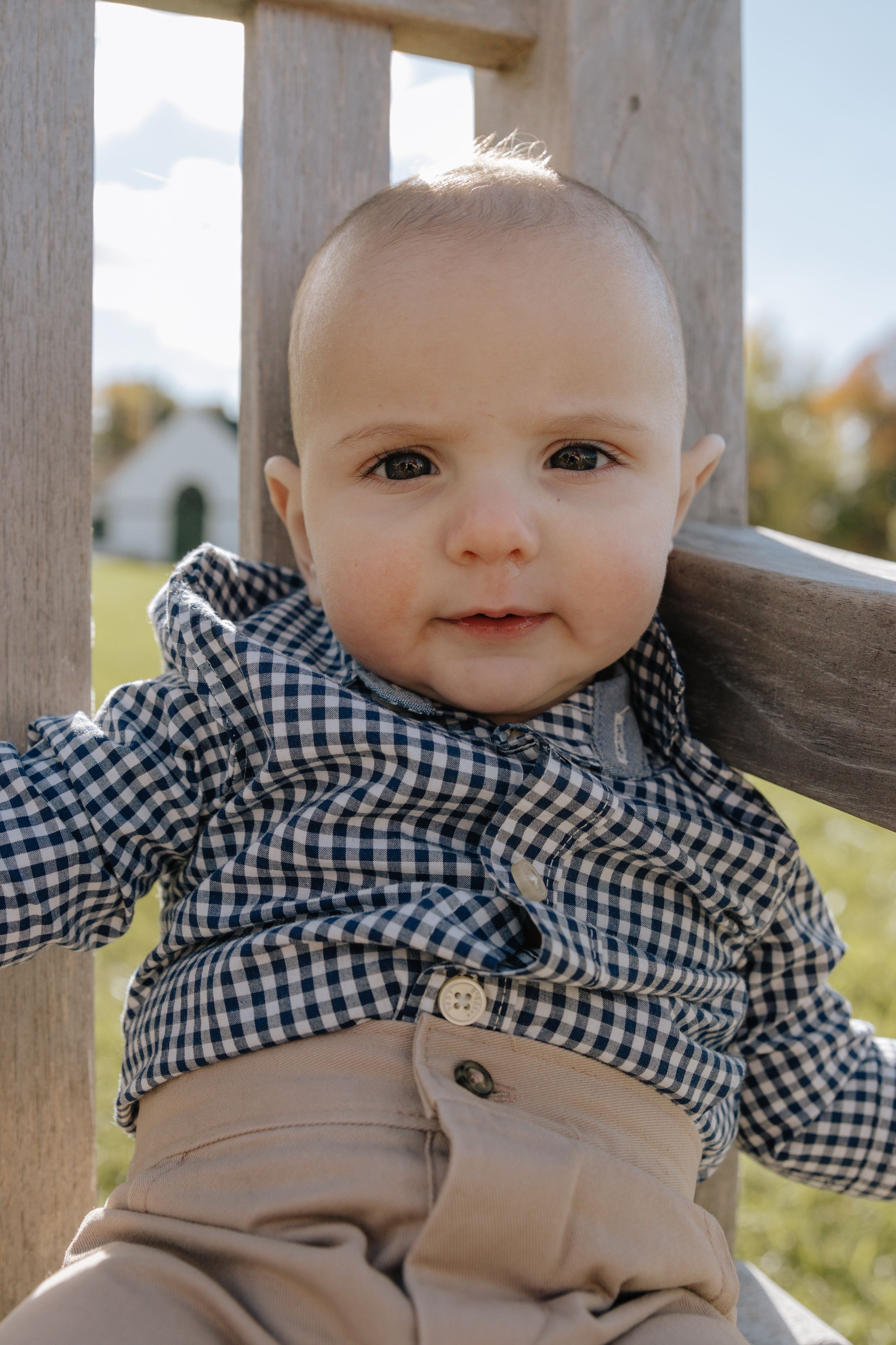 Sweet baby in checkered shirt smiling while sitting on wooden fence.