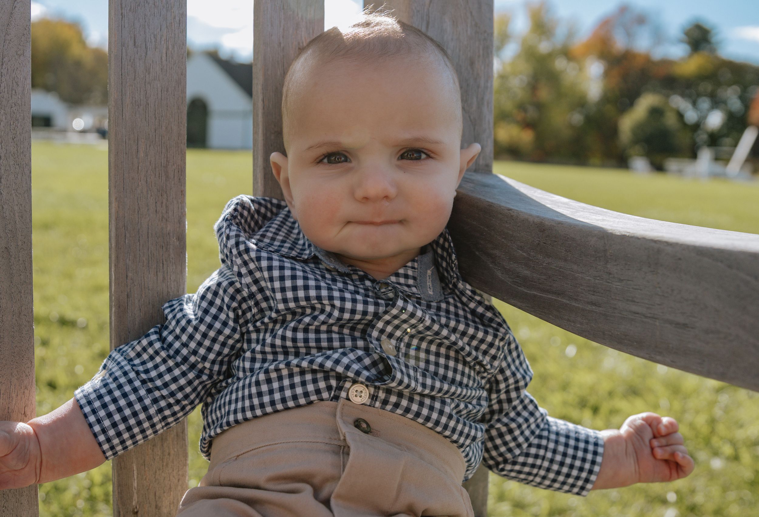 Baby in gingham shirt posing by white fence during outdoor photo session.