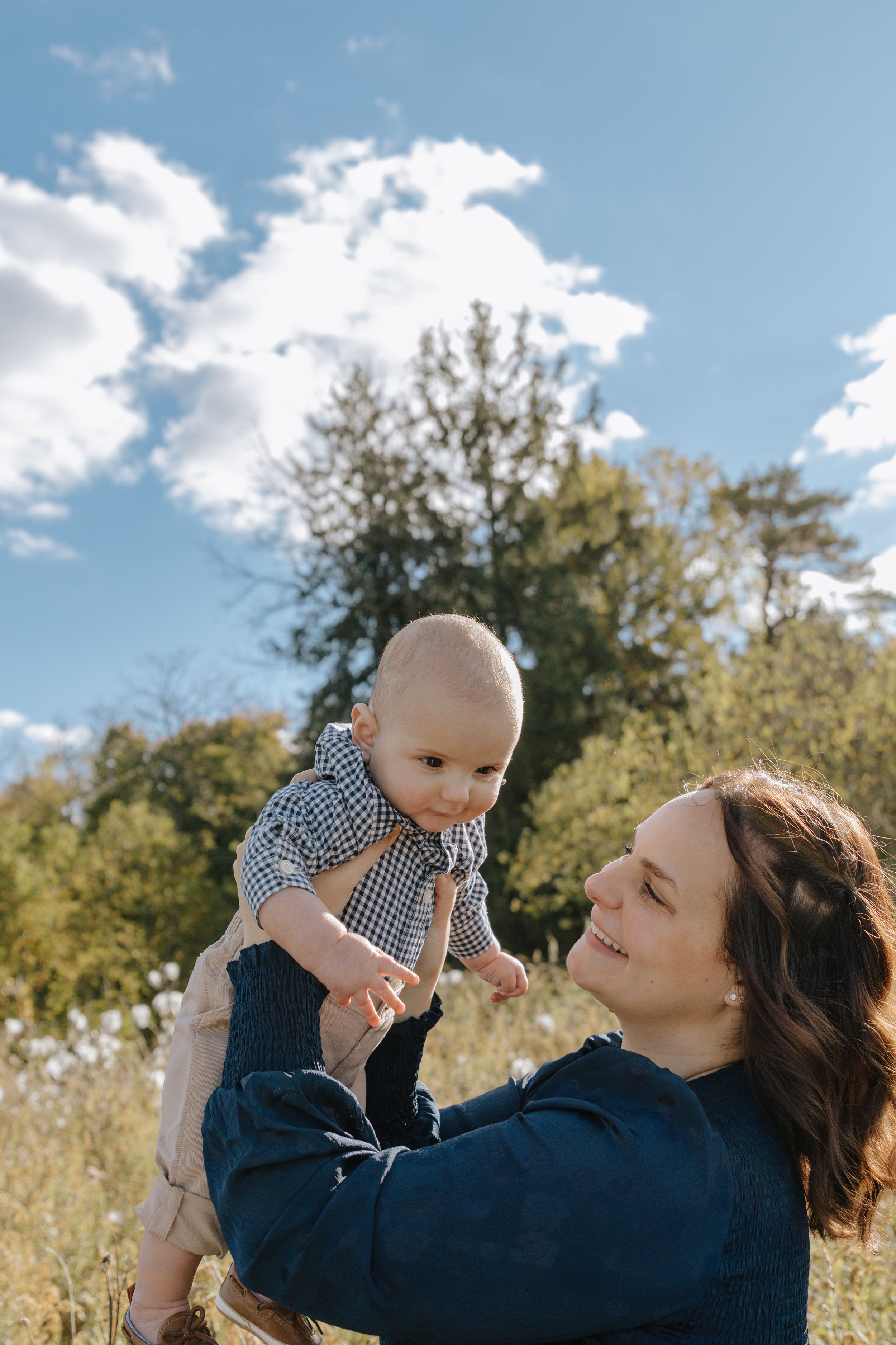 A tender outdoor moment between parent and baby against a blue sky and autumn trees.