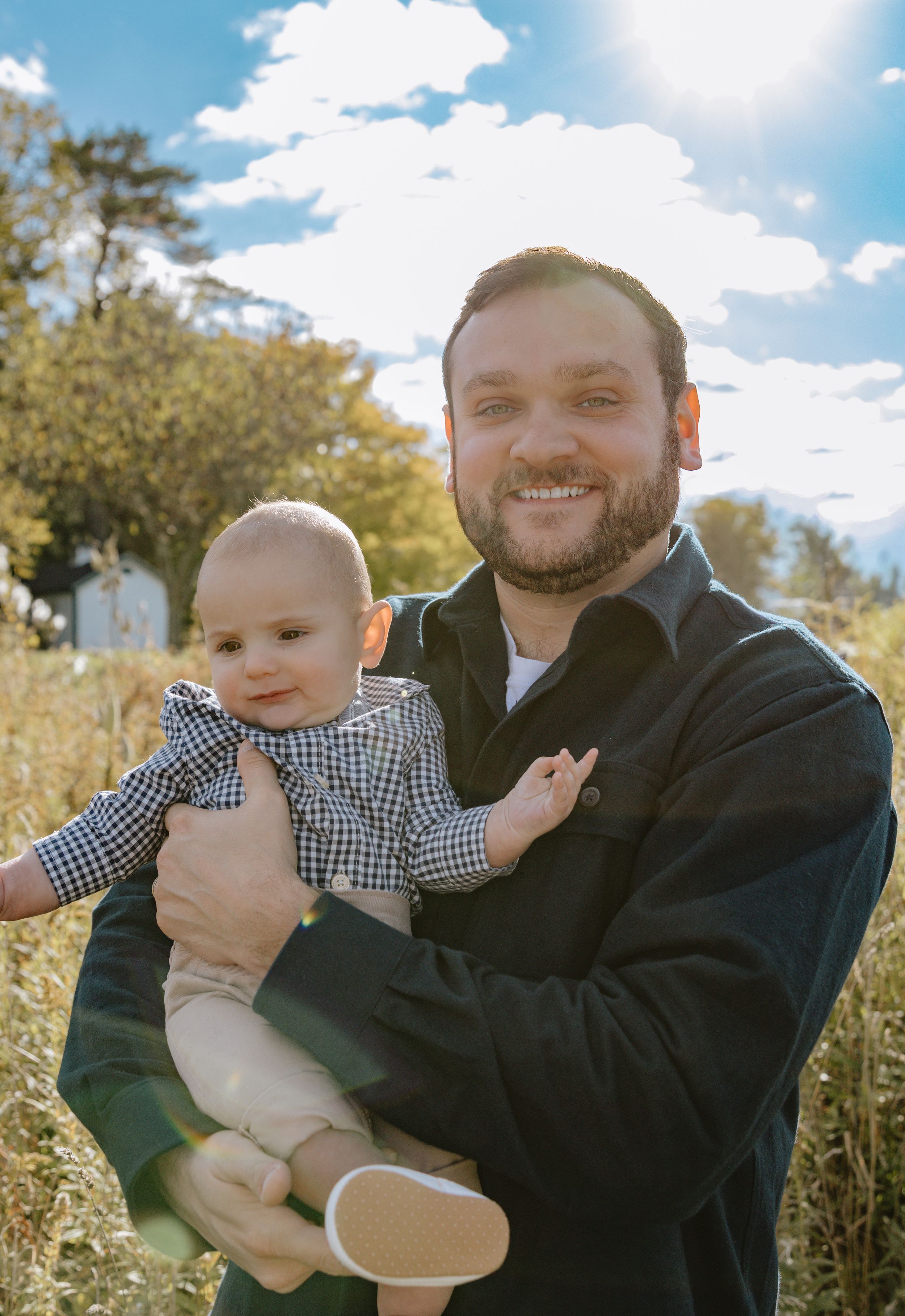 Joyful outdoor family portrait with baby wearing checkered shirt in natural setting.