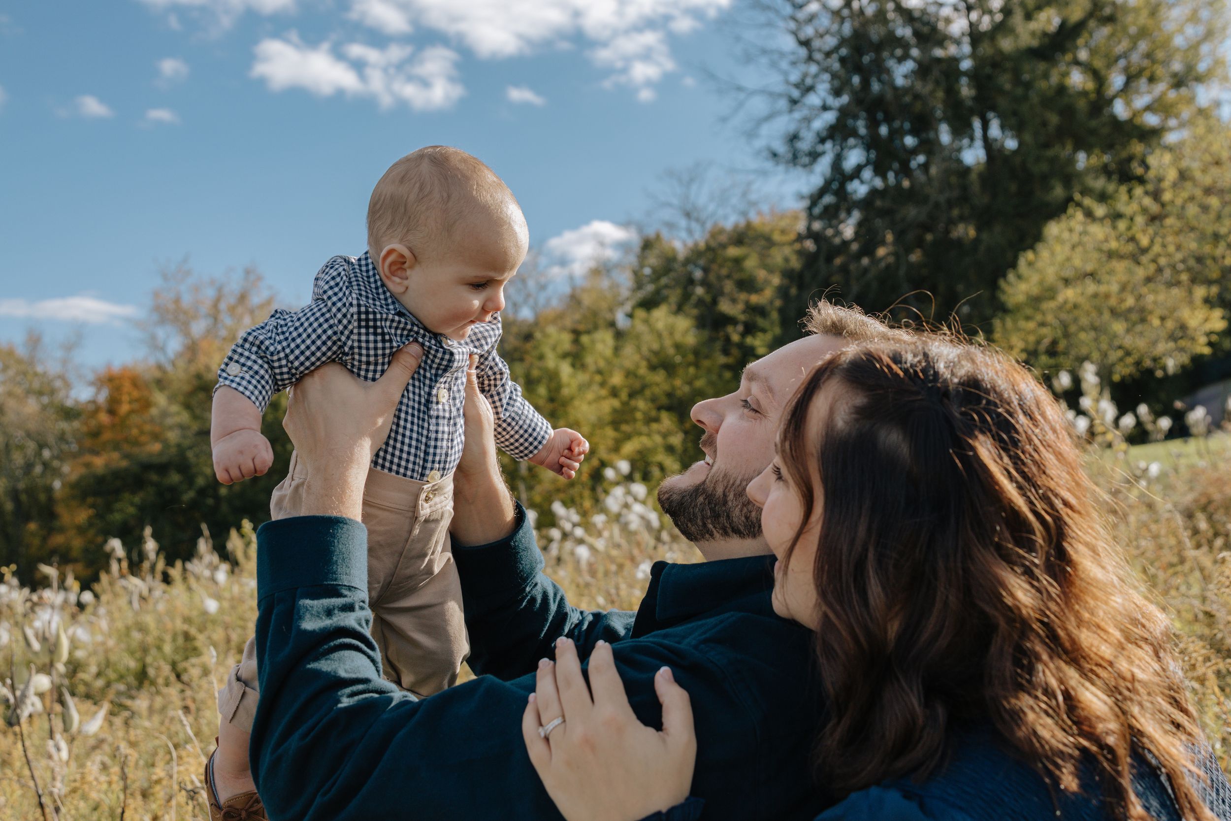 Family plays together outdoors lifting baby in sunny autumn field.