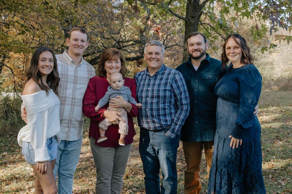 Family group stands together smiling in front of fall foliage.