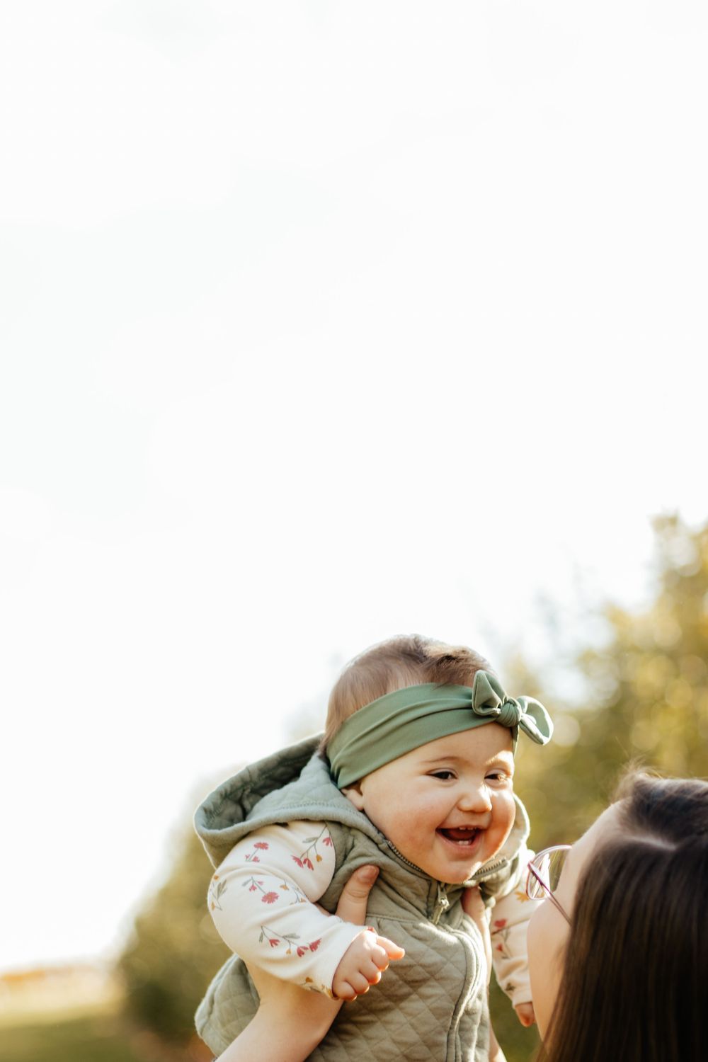 A joyful baby wearing a green headband is lifted high in the air during an outdoor photoshoot.