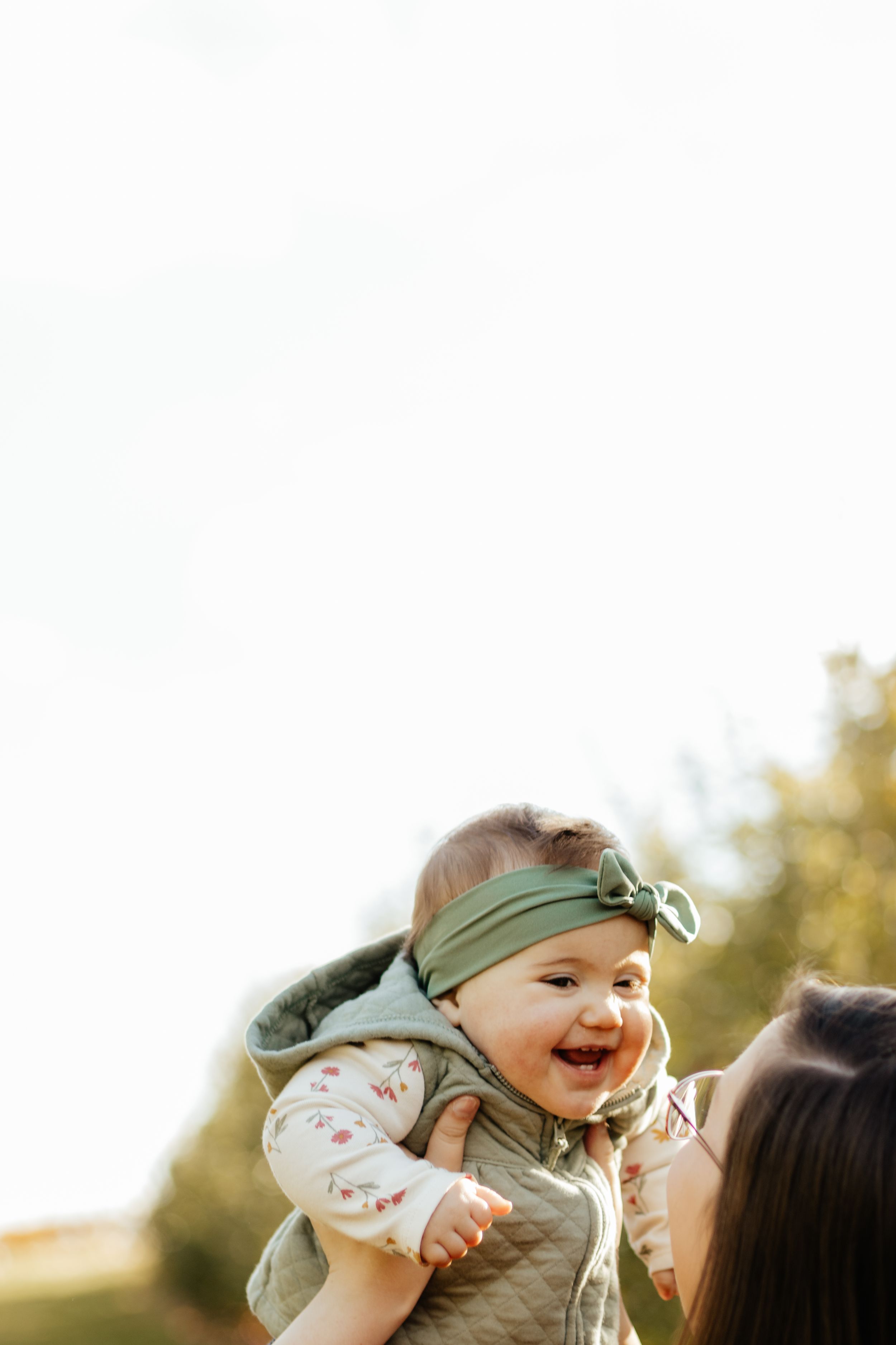 A joyful baby wearing a green headband is lifted high in the air during an outdoor photoshoot.