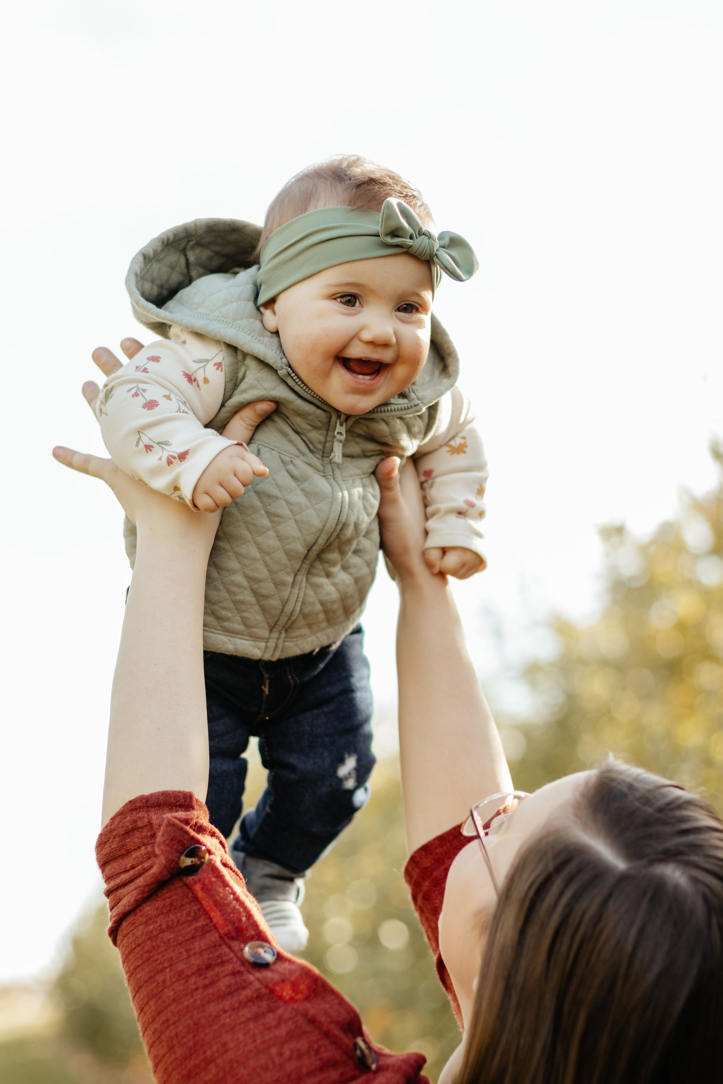 A laughing baby in a green hat and vest is lifted playfully during a sunny outdoor portrait session.