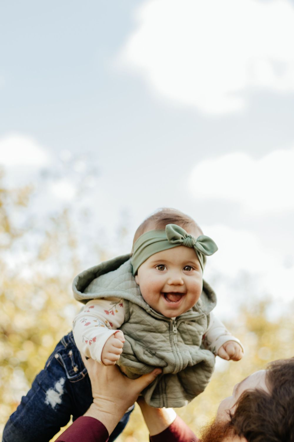 A baby wearing a sage green headband and vest is lifted joyfully into the air against a bright sky.