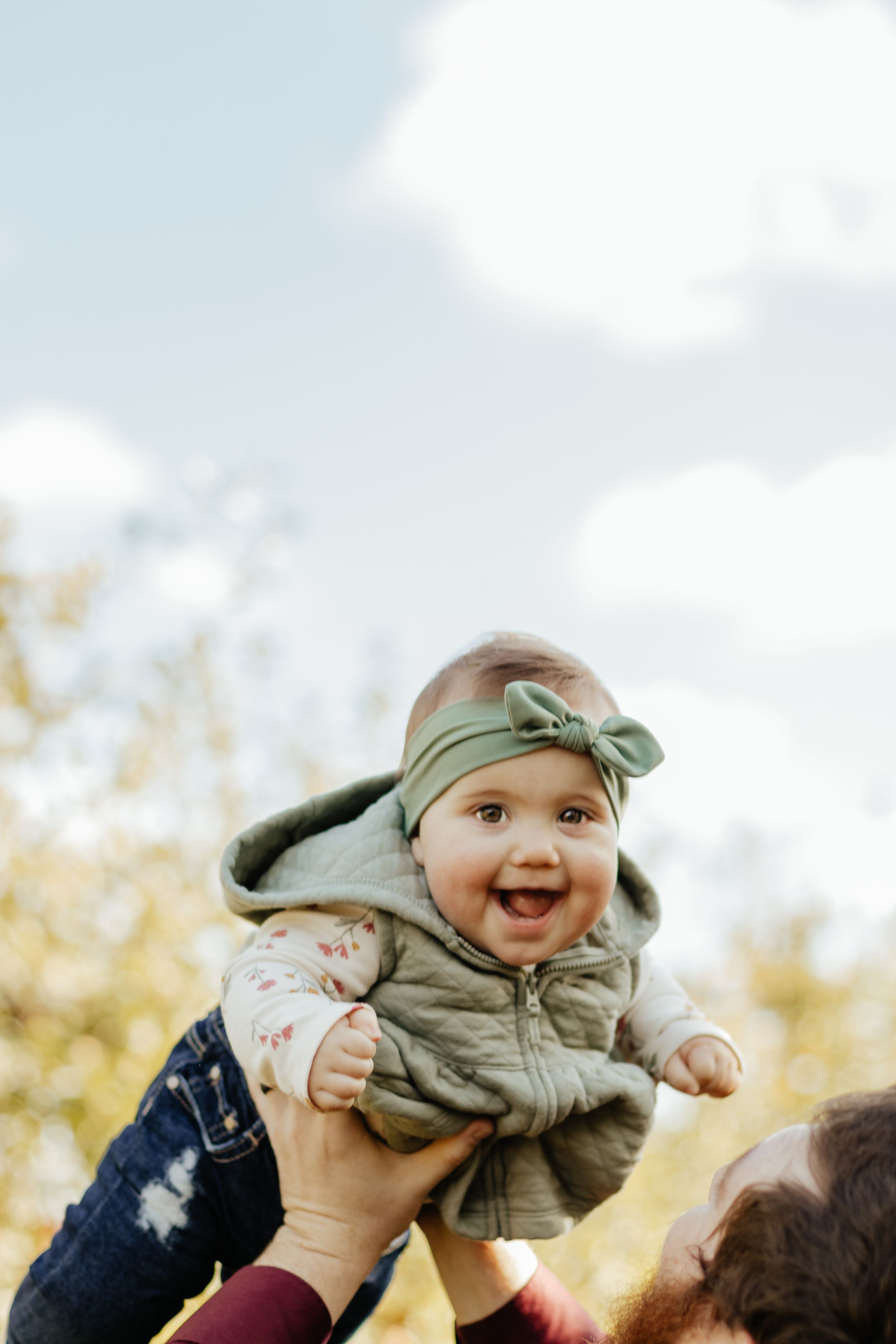 A baby wearing a sage green headband and vest is lifted joyfully into the air against a bright sky.