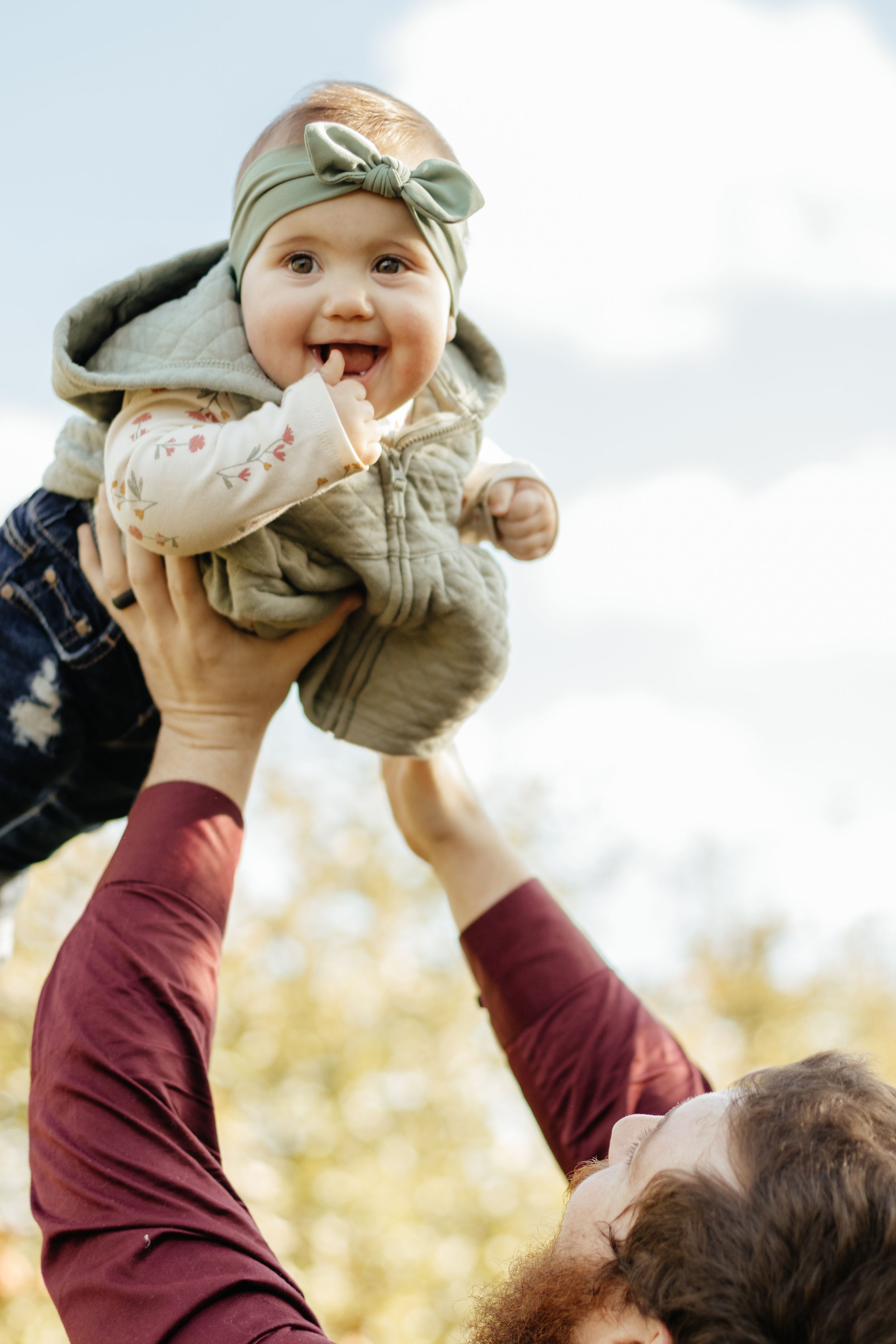A laughing baby in green clothing is held up high by someone in a burgundy shirt.