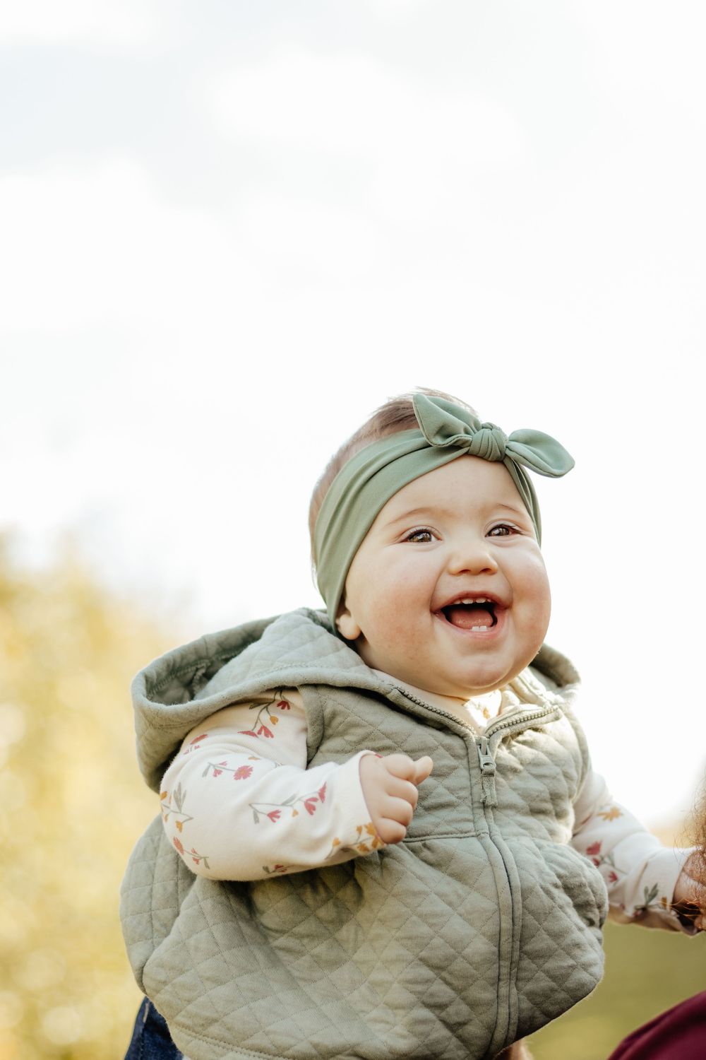 A cheerful baby in a quilted vest and bow headband smiles brightly outdoors.