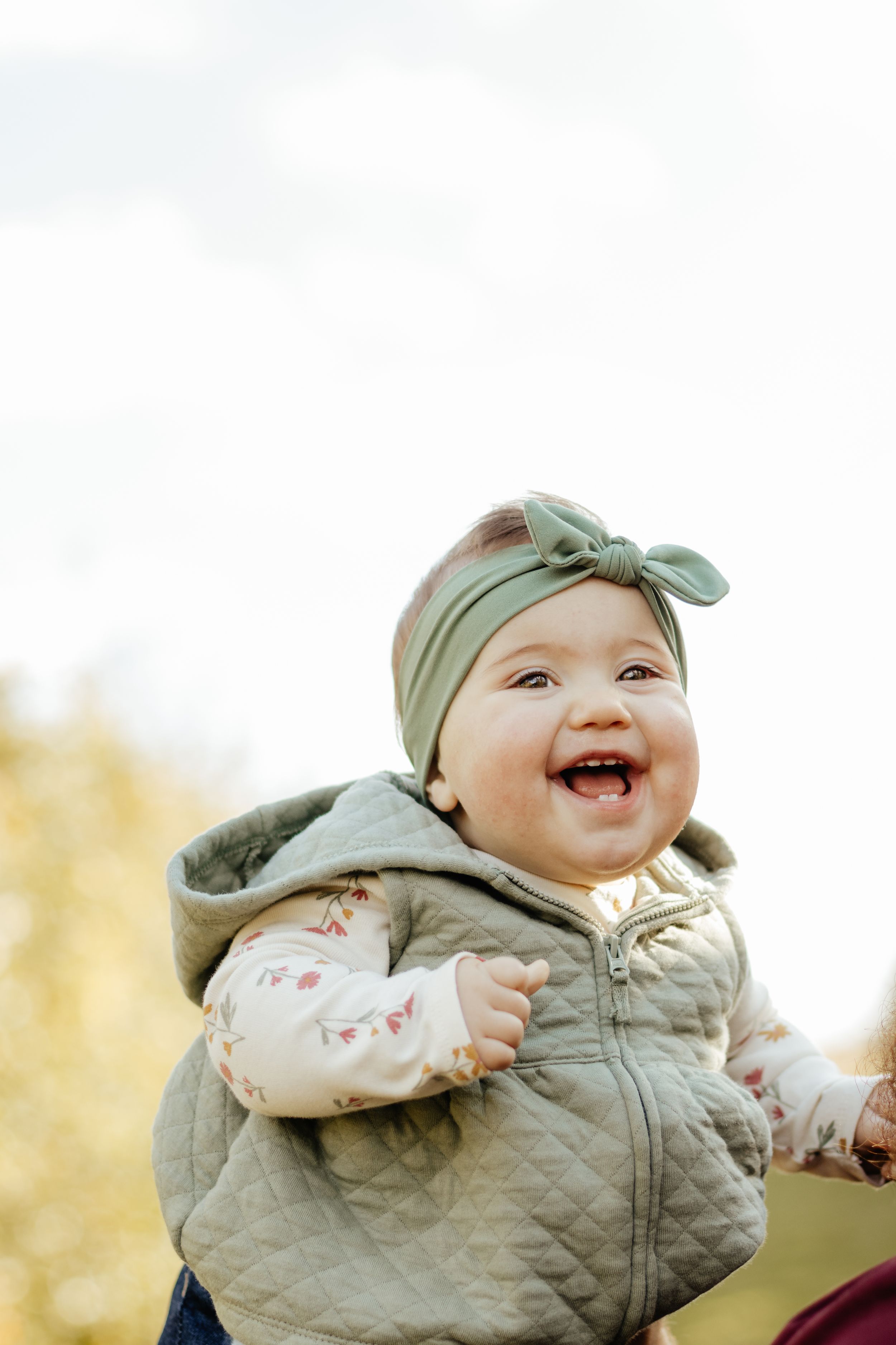 A cheerful baby in a quilted vest and bow headband smiles brightly outdoors.