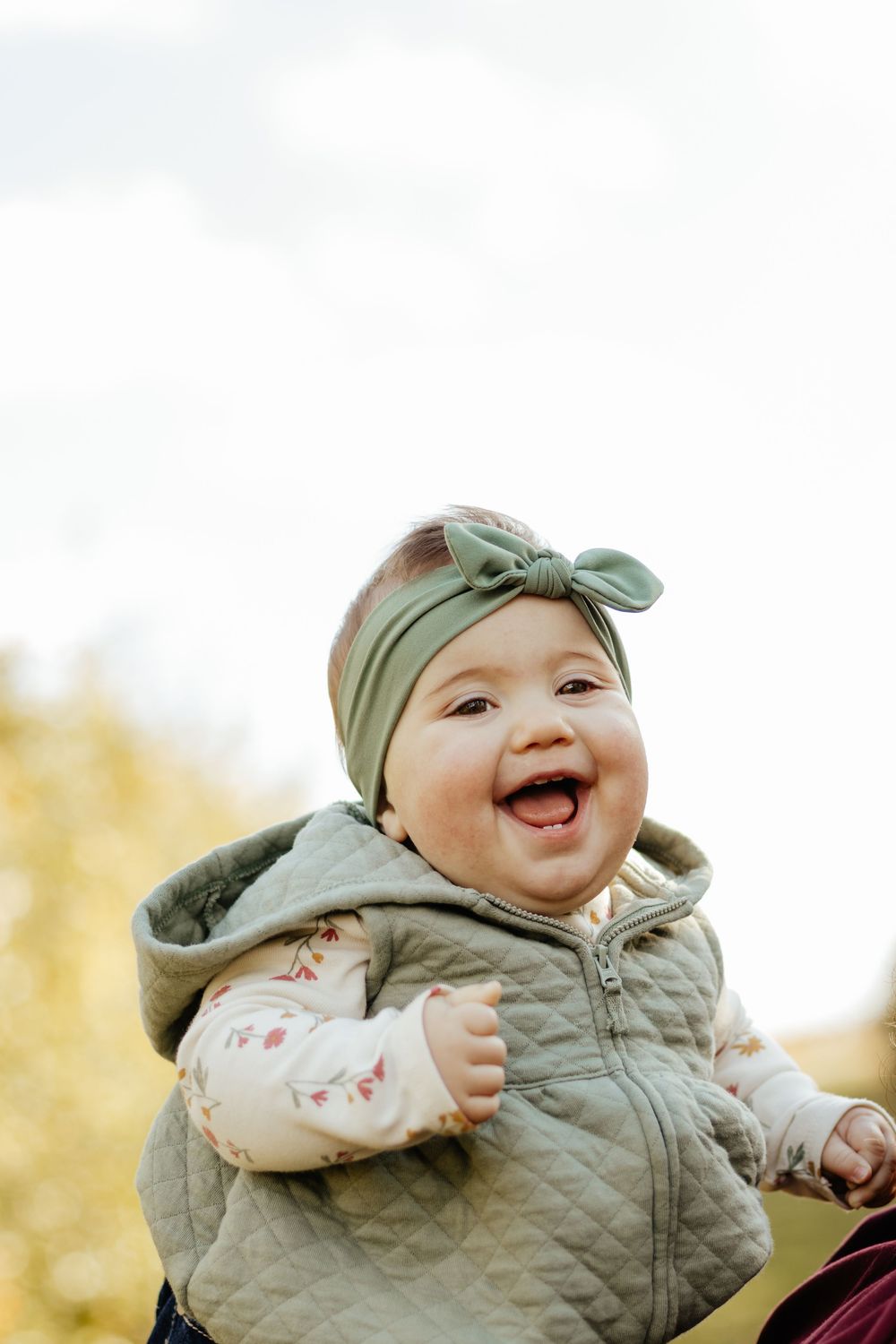 A happy infant wearing a sage green outfit laughs while being lifted up during playtime.
