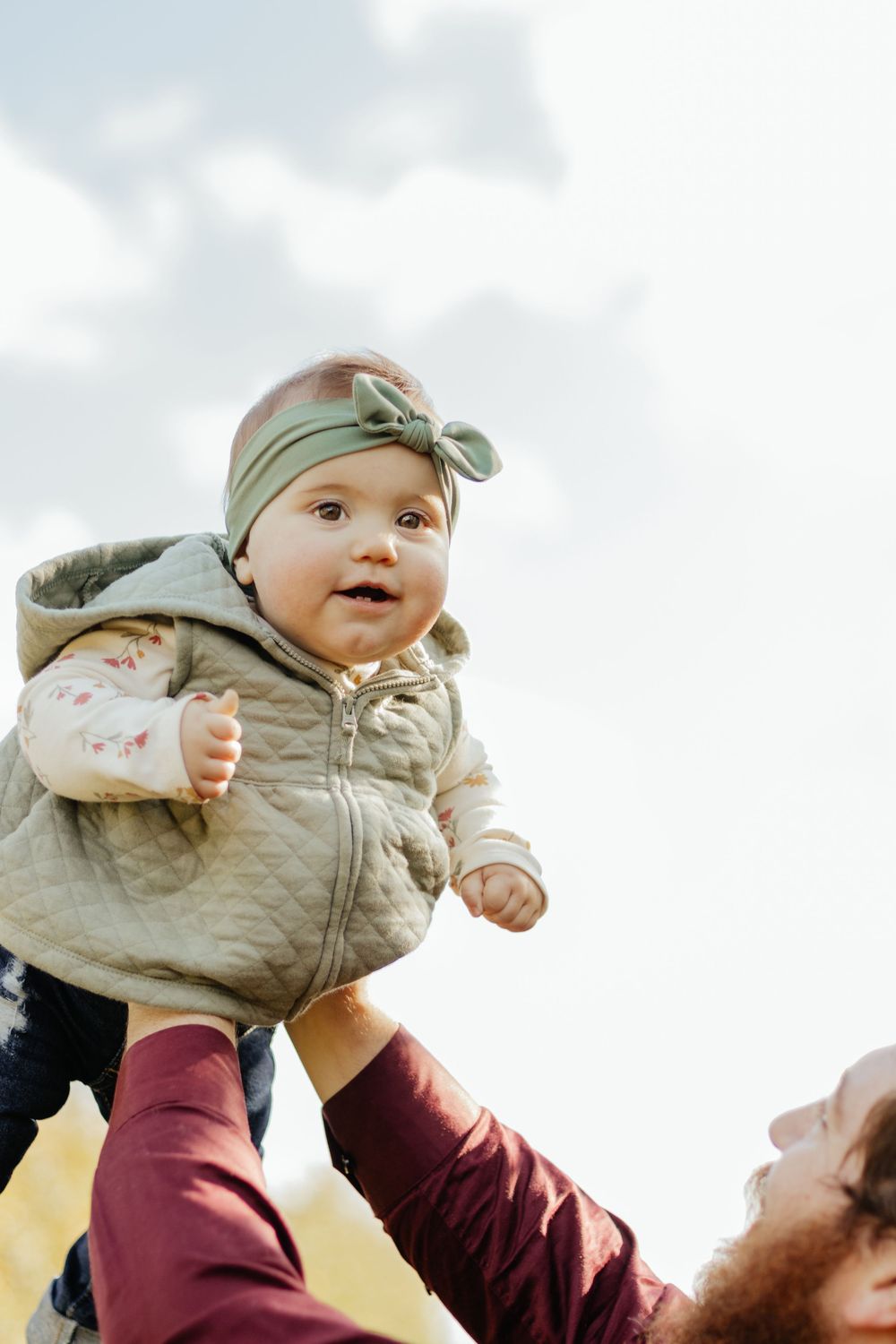 A baby in a quilted vest and matching headband is lifted up against a cloudy sky.