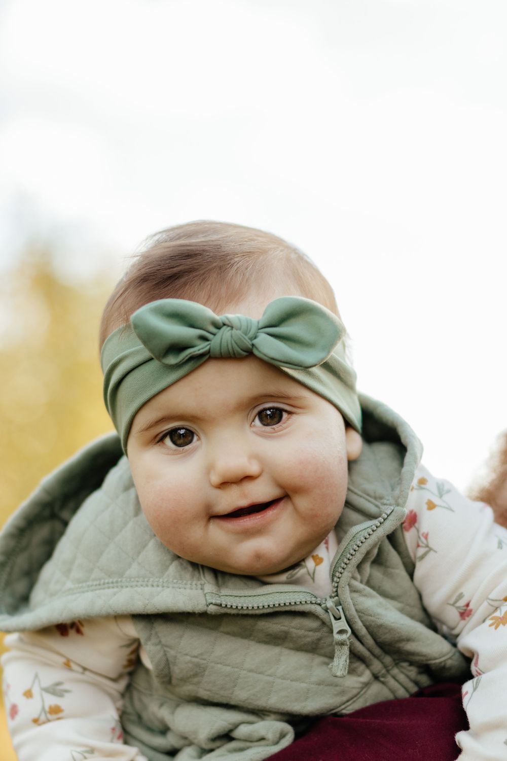 Close-up of an infant wearing a sage green bow headband and quilted vest.