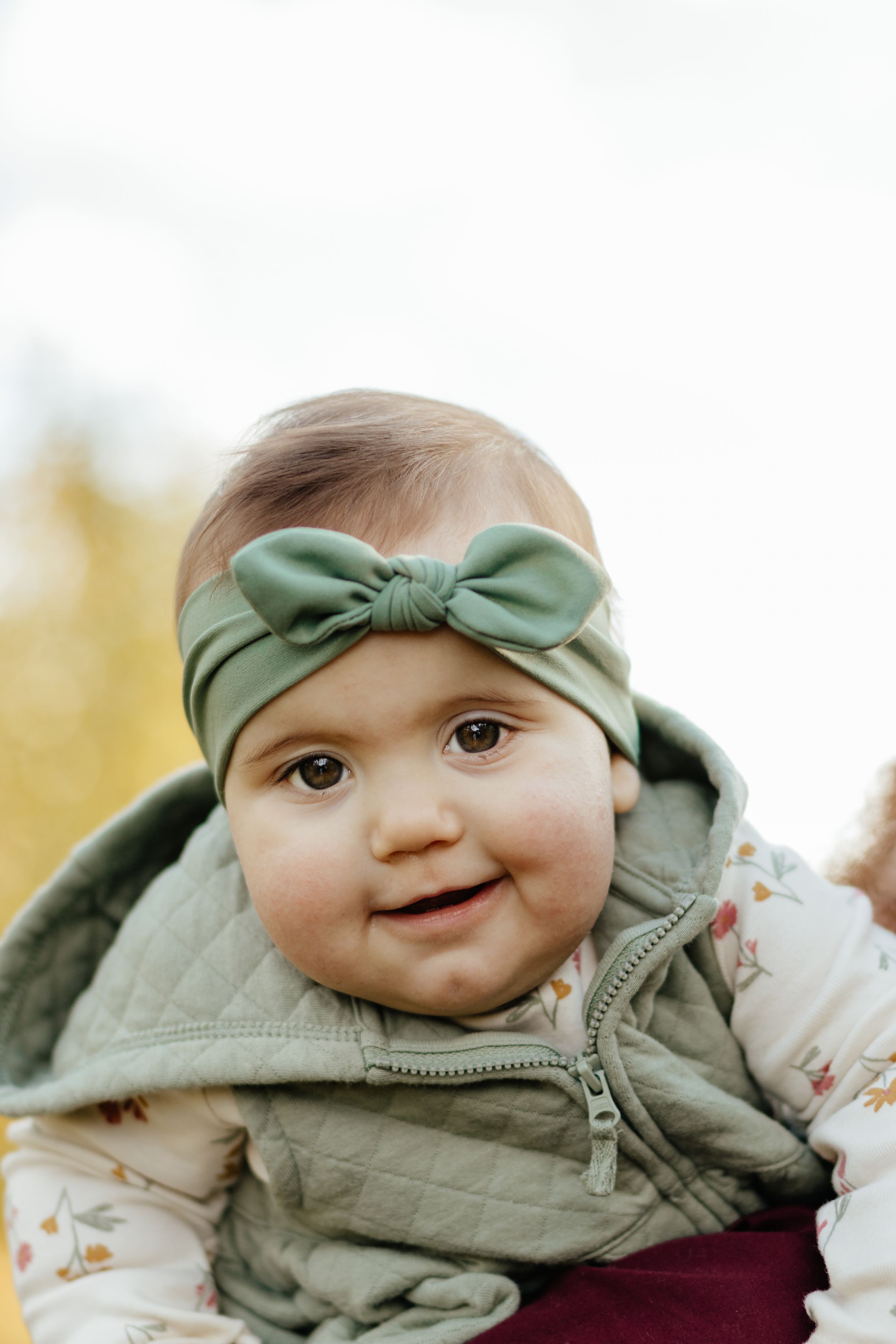 Close-up of an infant wearing a sage green bow headband and quilted vest.