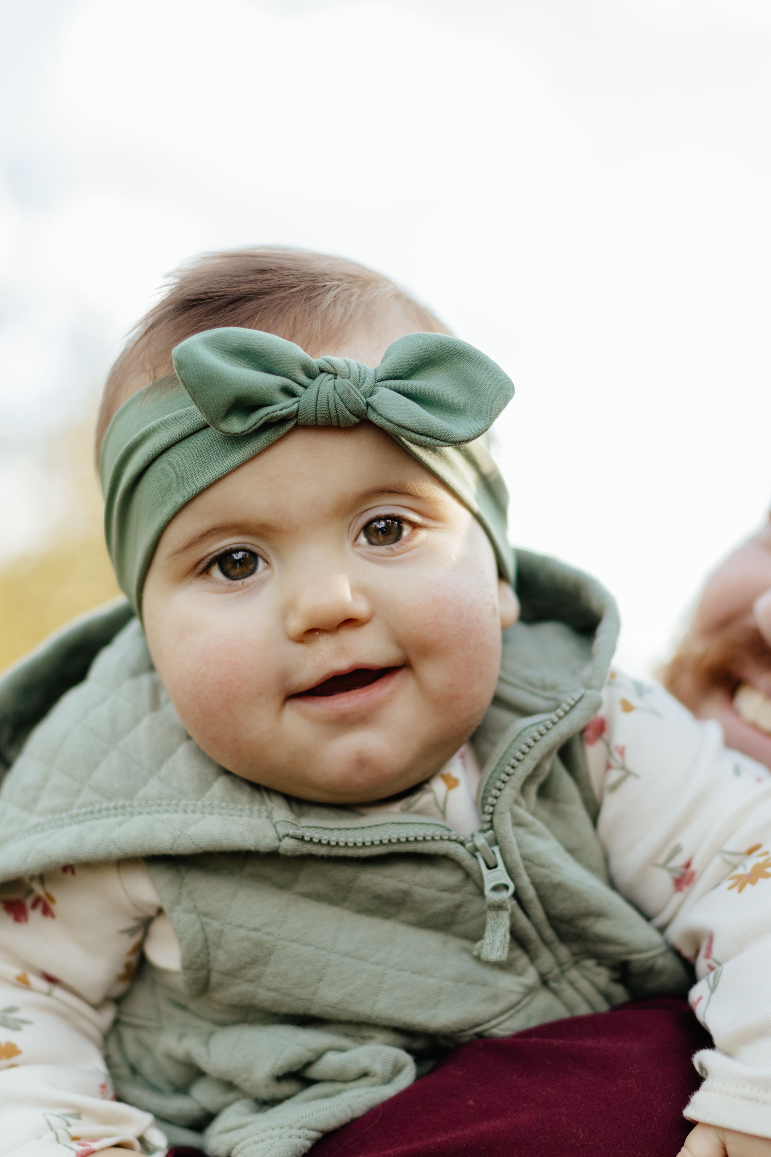Close portrait of a baby wearing a mint green bow and quilted vest smiling at the camera.