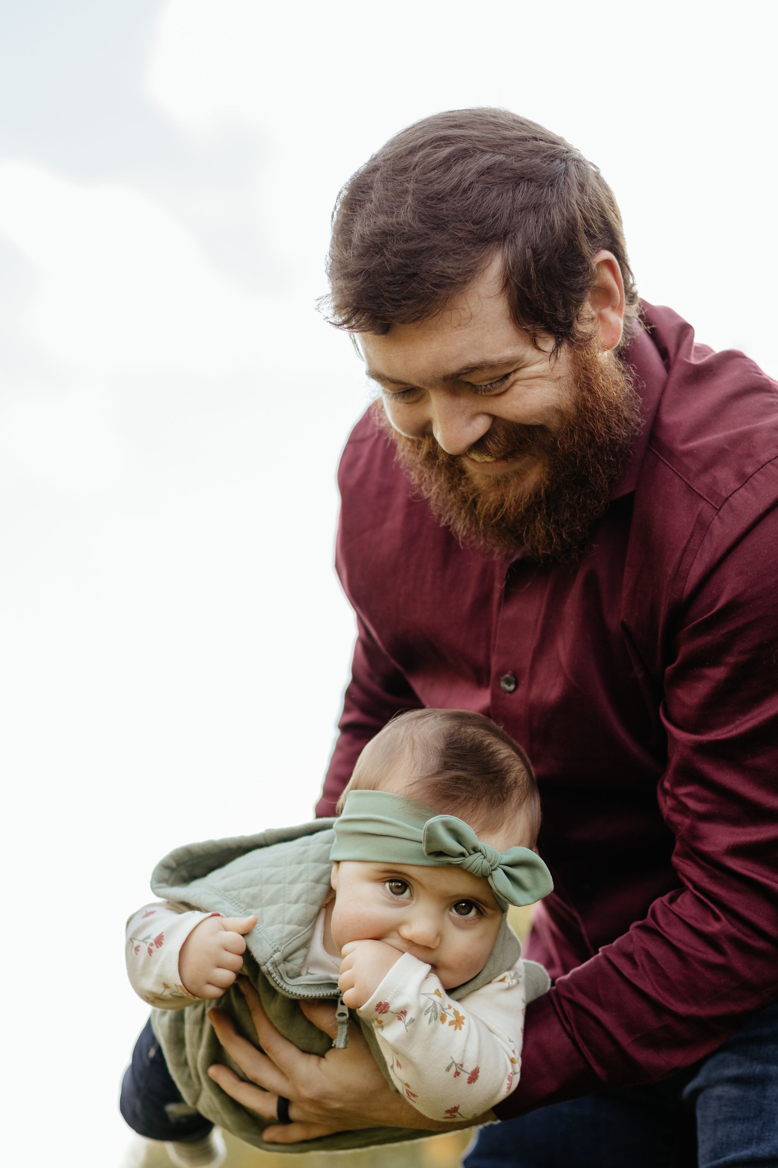 A bearded person in burgundy holds a smiling baby wearing green outdoors.