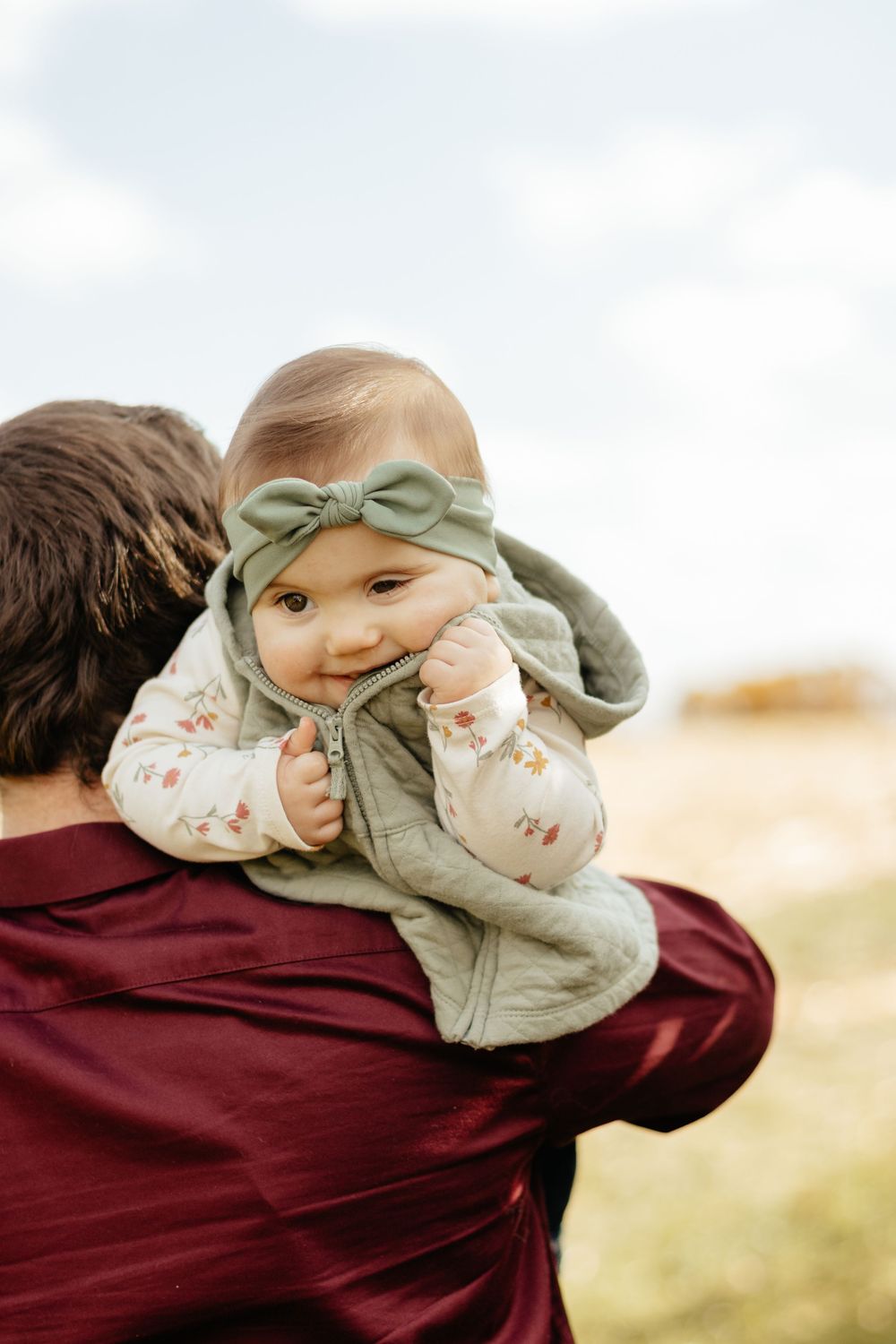 Baby wearing mint green headband and floral outfit being held during outdoor photoshoot.
