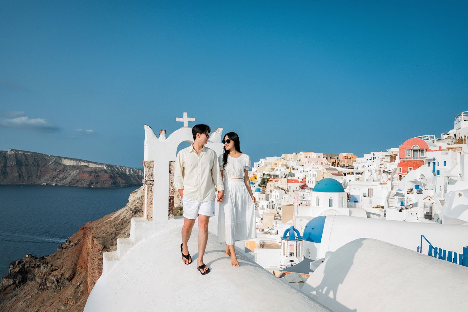 A romantic couple walks through the iconic white buildings and blue domes of Santorini, Greece.