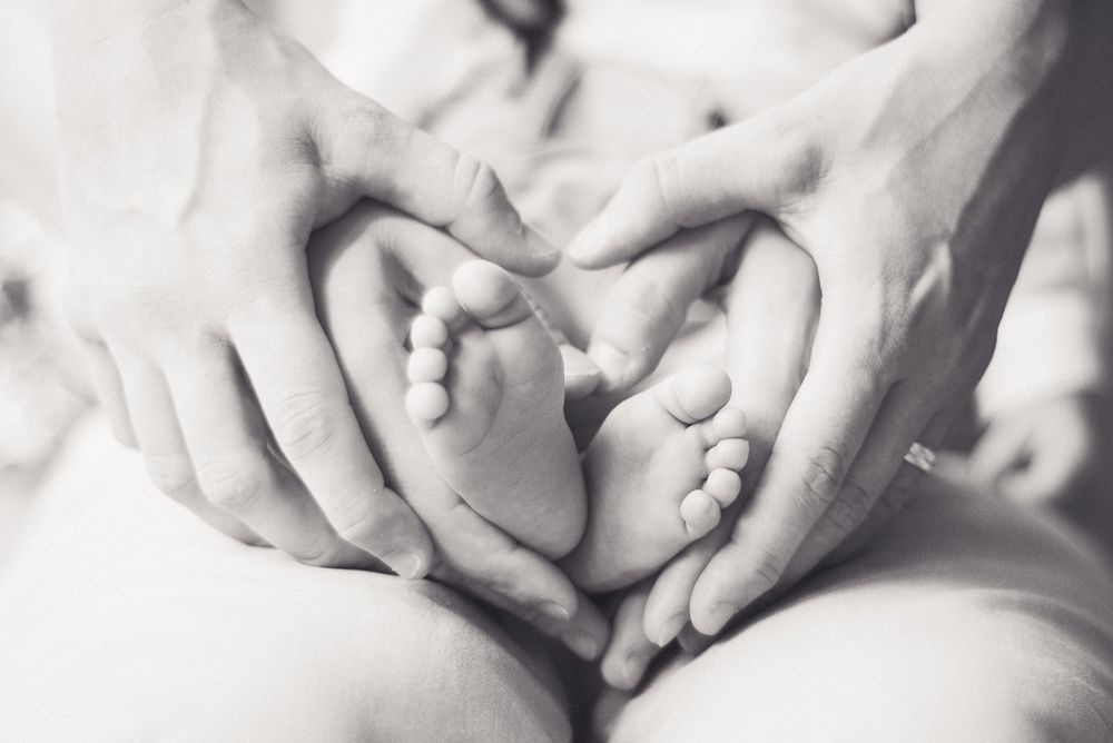 Détail en noir et blanc de mains formant un cœur autour de pieds de bébé, séance naissance intime et artistique.