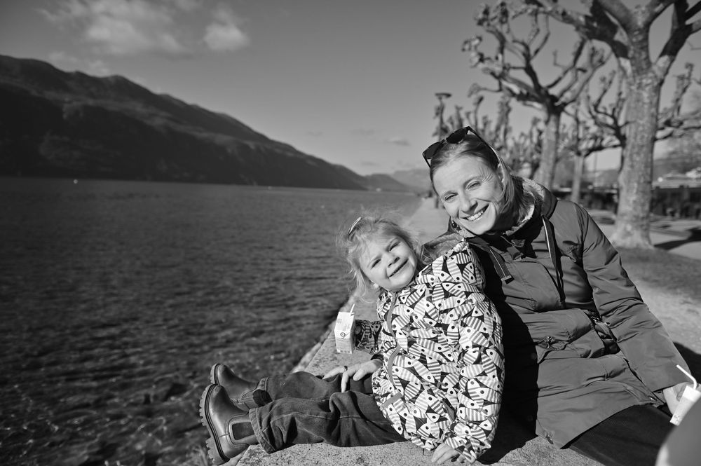Parent et enfant assis au bord d’un lac entouré de montagnes, portrait naturel en noir et blanc en extérieur.