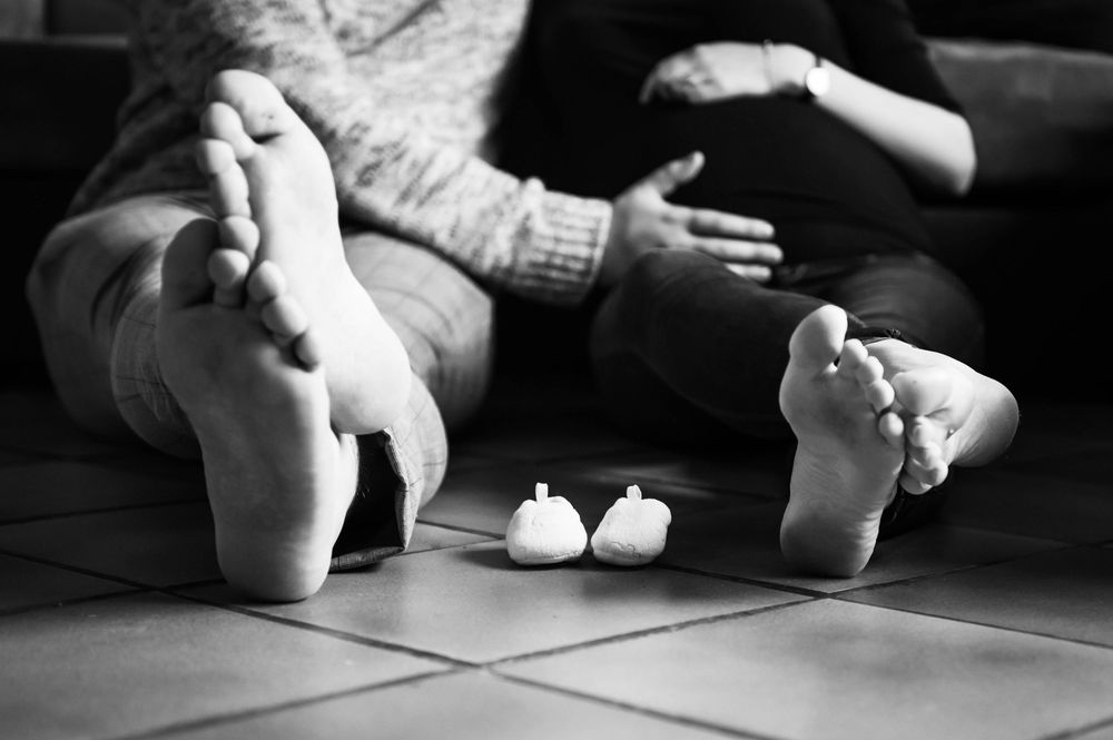 Détail en noir et blanc des pieds et mains d’un bébé avec parents en arrière-plan, séance grossesse intimiste en Bretagne.