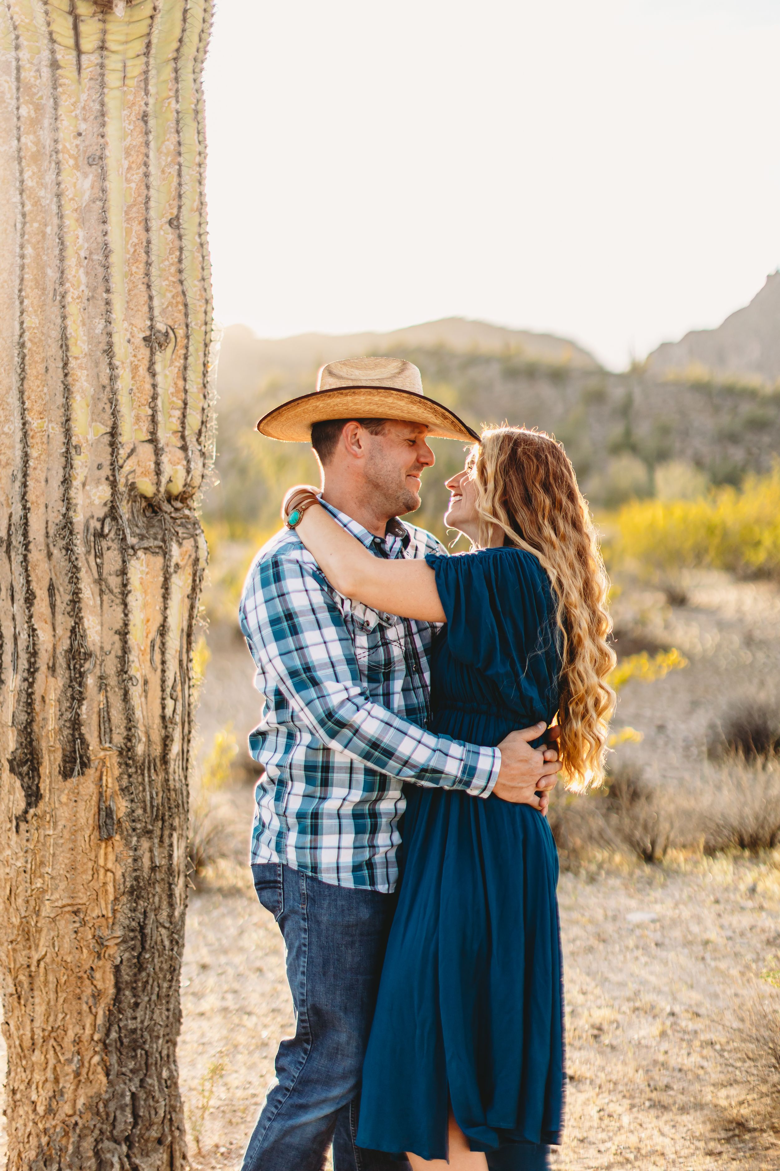 Couple poses intimately among tall saguaro cacti in desert setting at sunset.