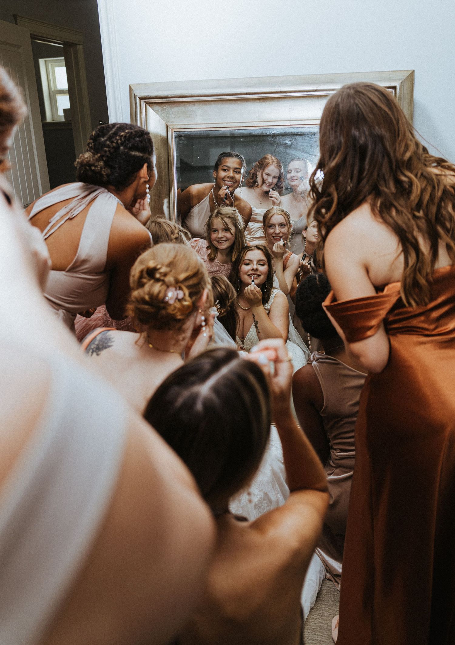 A bride and all of her bridesmaids lean into the mirror as she puts lip gloss on