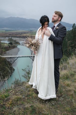 A couple in wedding attire embraces on a scenic overlook with turquoise river waters and mountains in the background.