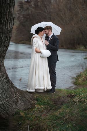 A romantic winter wedding couple shares an intimate moment under a white umbrella by a frozen lake.