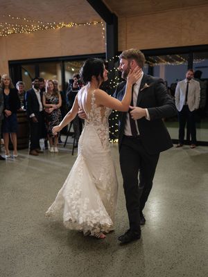 A couple shares their first dance at a wedding reception with string lights and guests watching in the background.