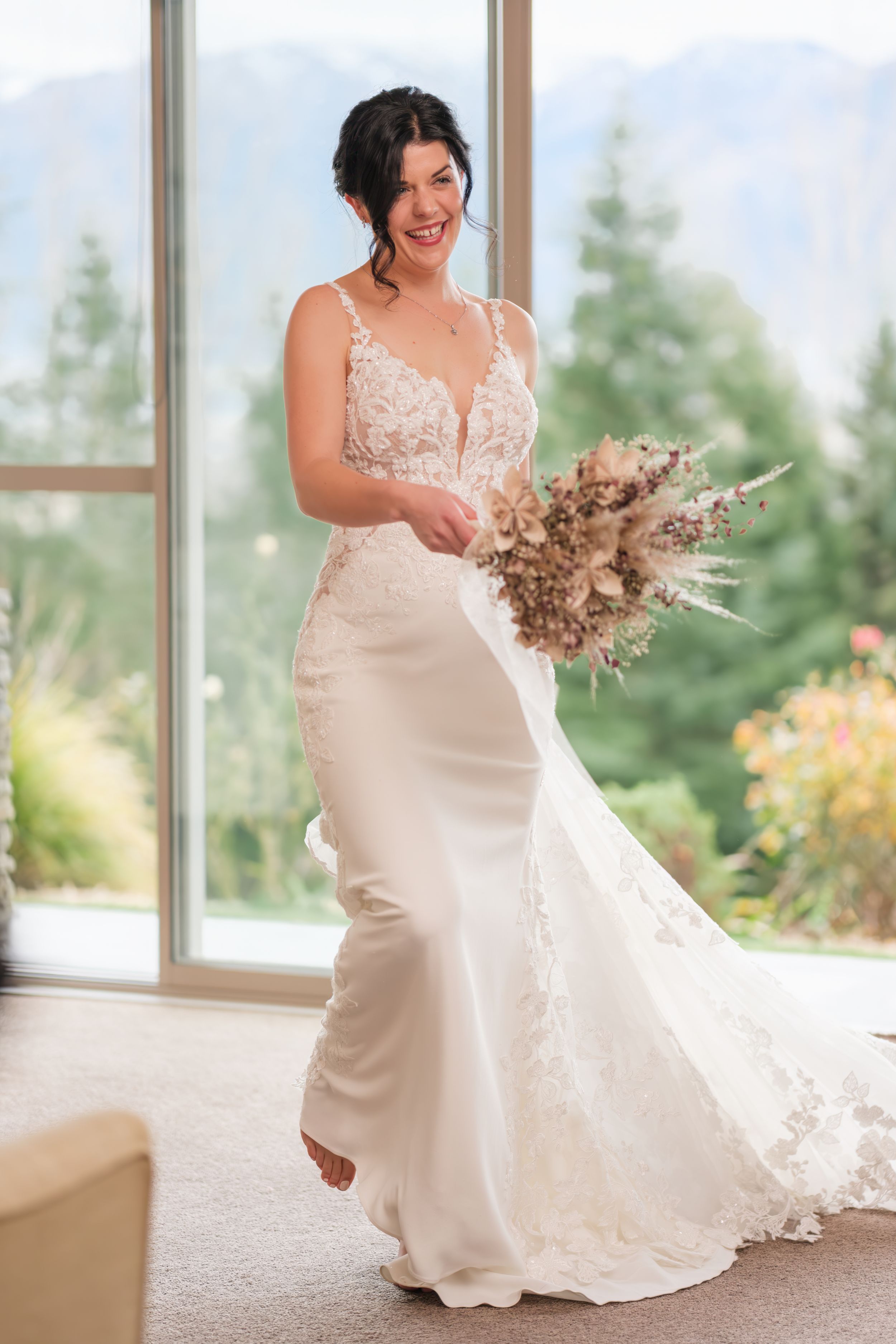 A bride in a white satin wedding dress twirls and dances while holding a dried floral bouquet in a sunlit room.