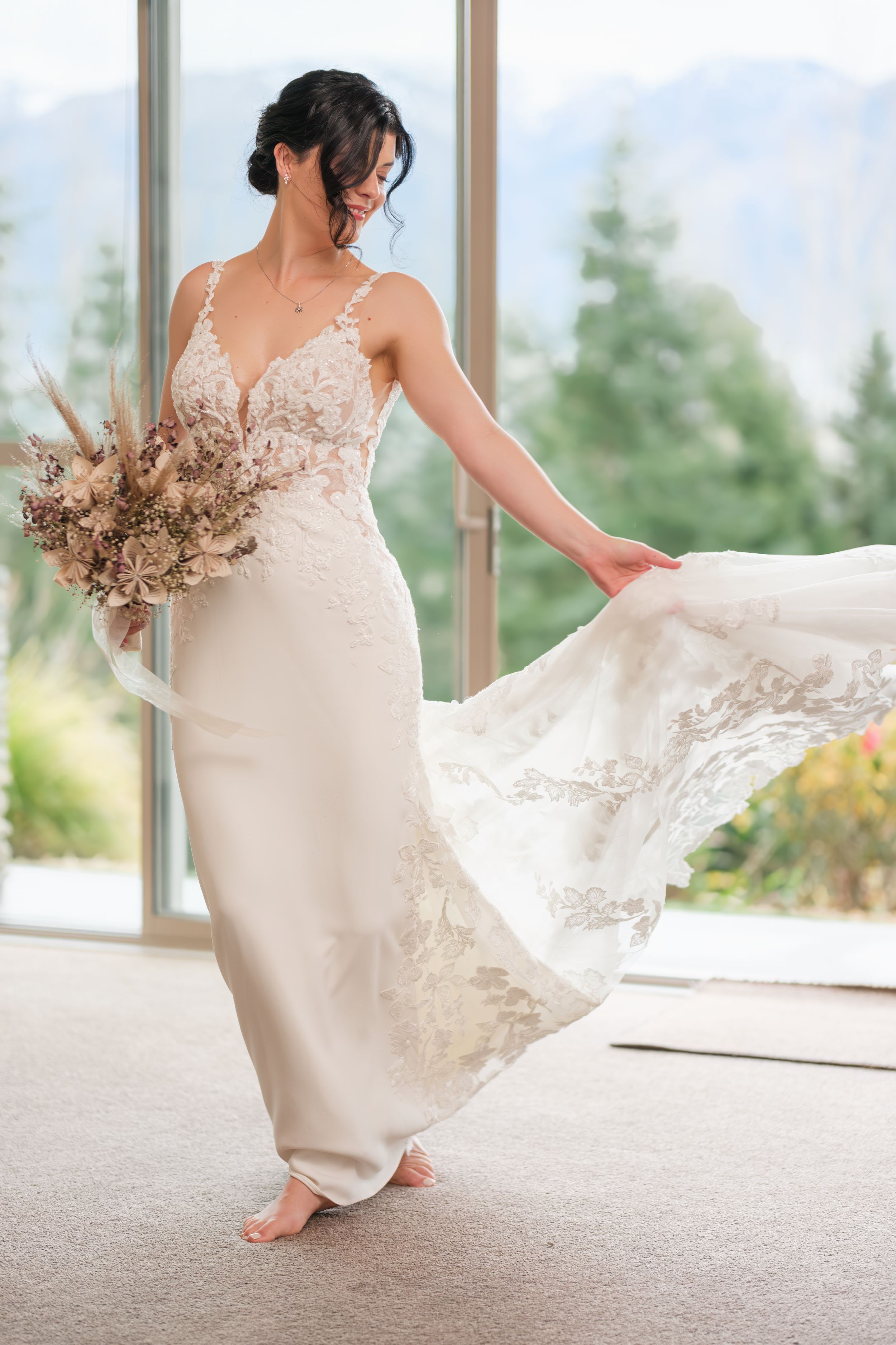 Elegant bride in lace wedding dress twirls barefoot while holding dried floral bouquet near bright window.
