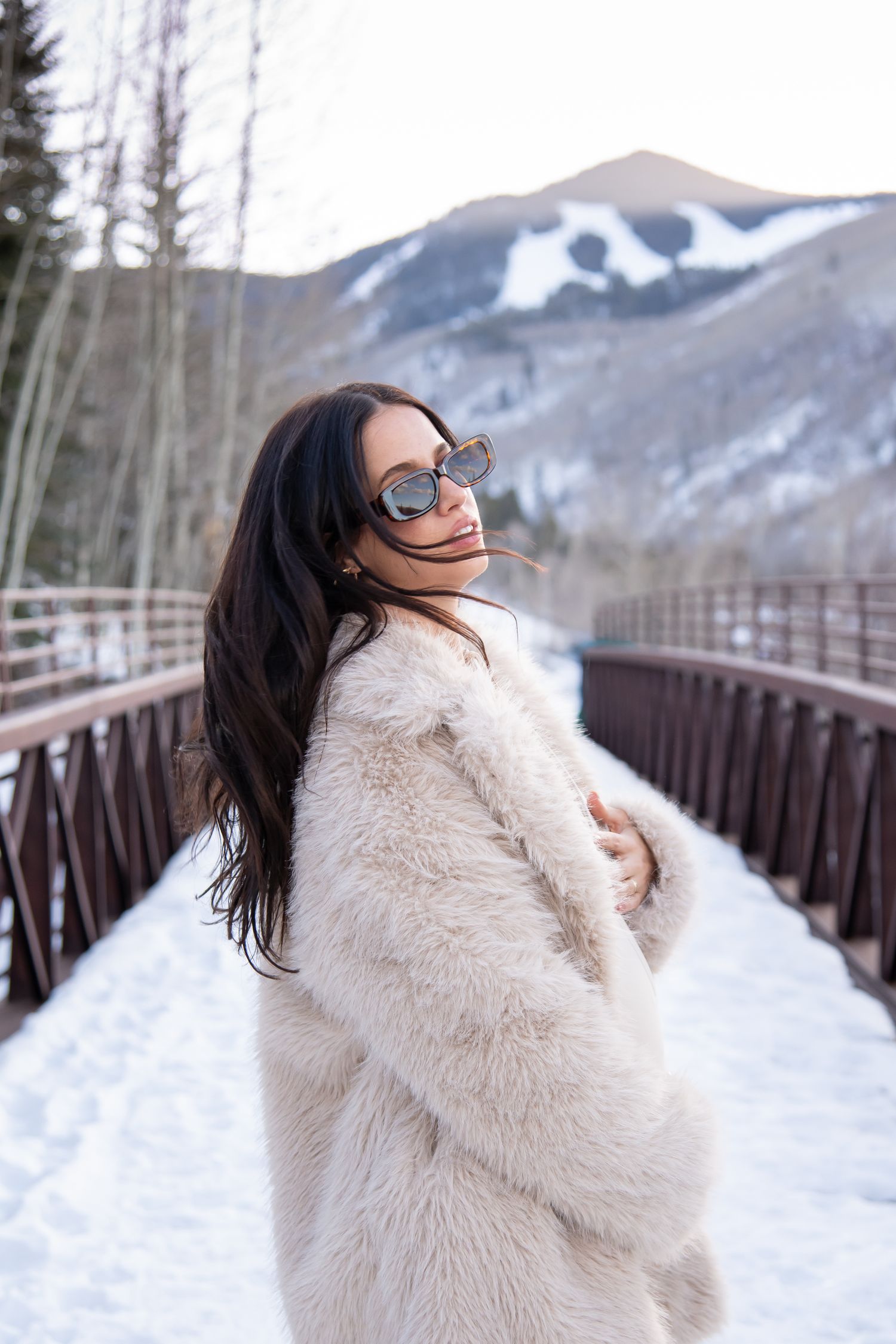 Candid editorial style photo of a stylish woman with beige fur coat with snowy mountains during her winter maternity photo session in Vail