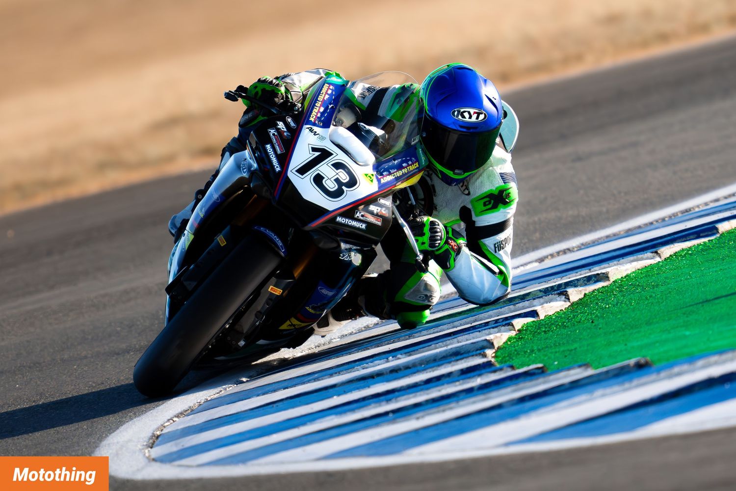Motorcycle racers lean into a turn on a race track with blue and green curbing visible at the edge.