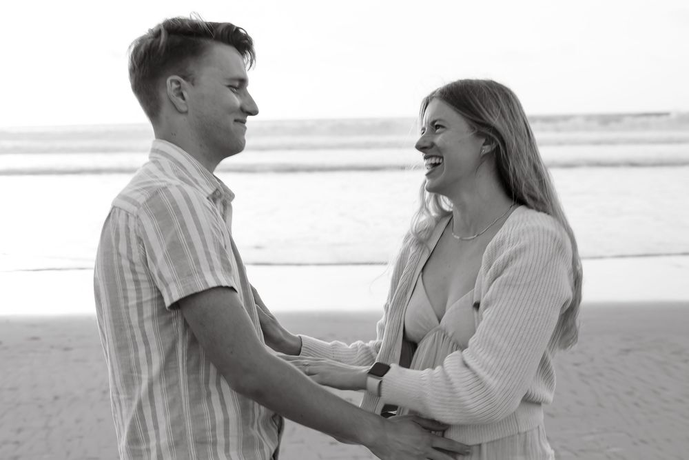 Black and white photo of laughing couple holding hands facing each other on beach.