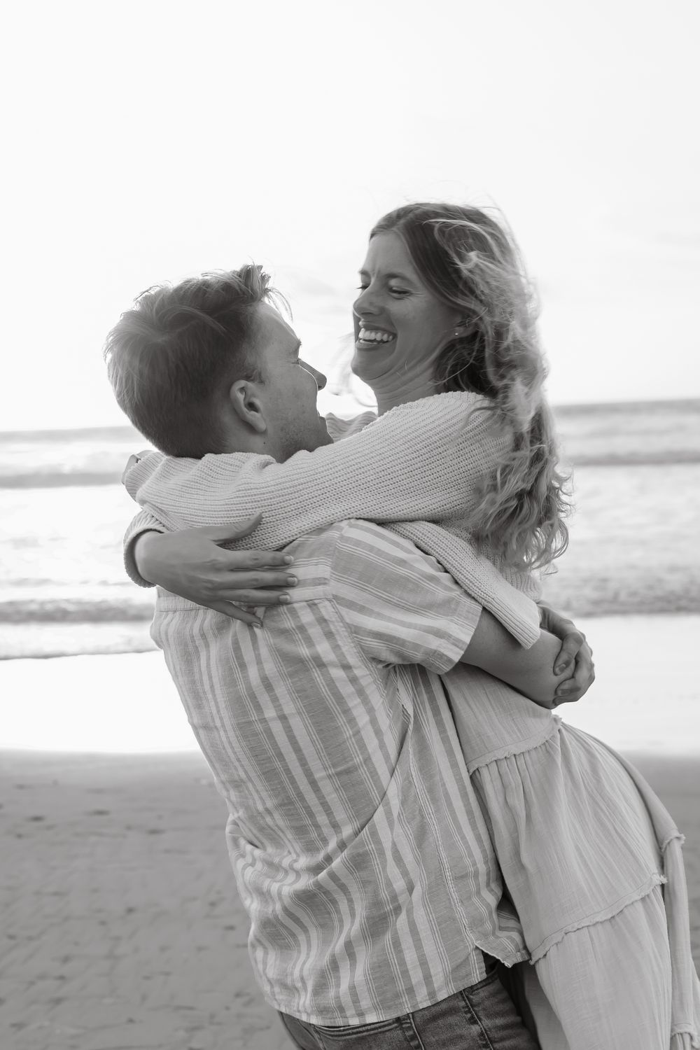 Black and white photo of joyful couple hugging and laughing together on sandy beach.