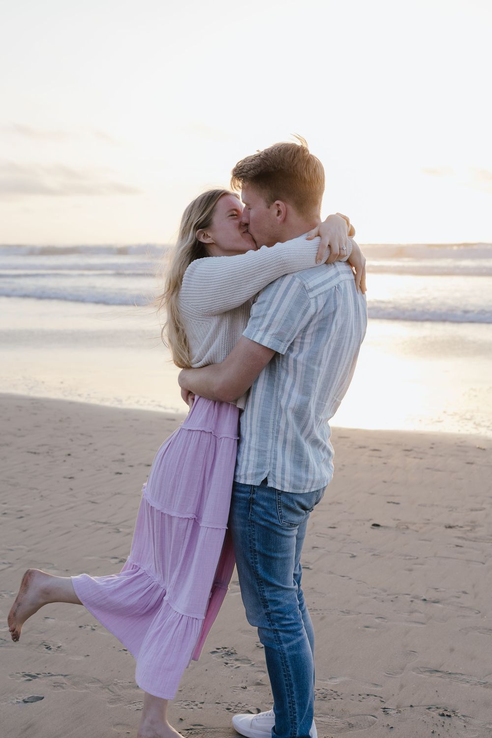 Couple shares a tender kiss while embracing on the beach during a colorful sunset.